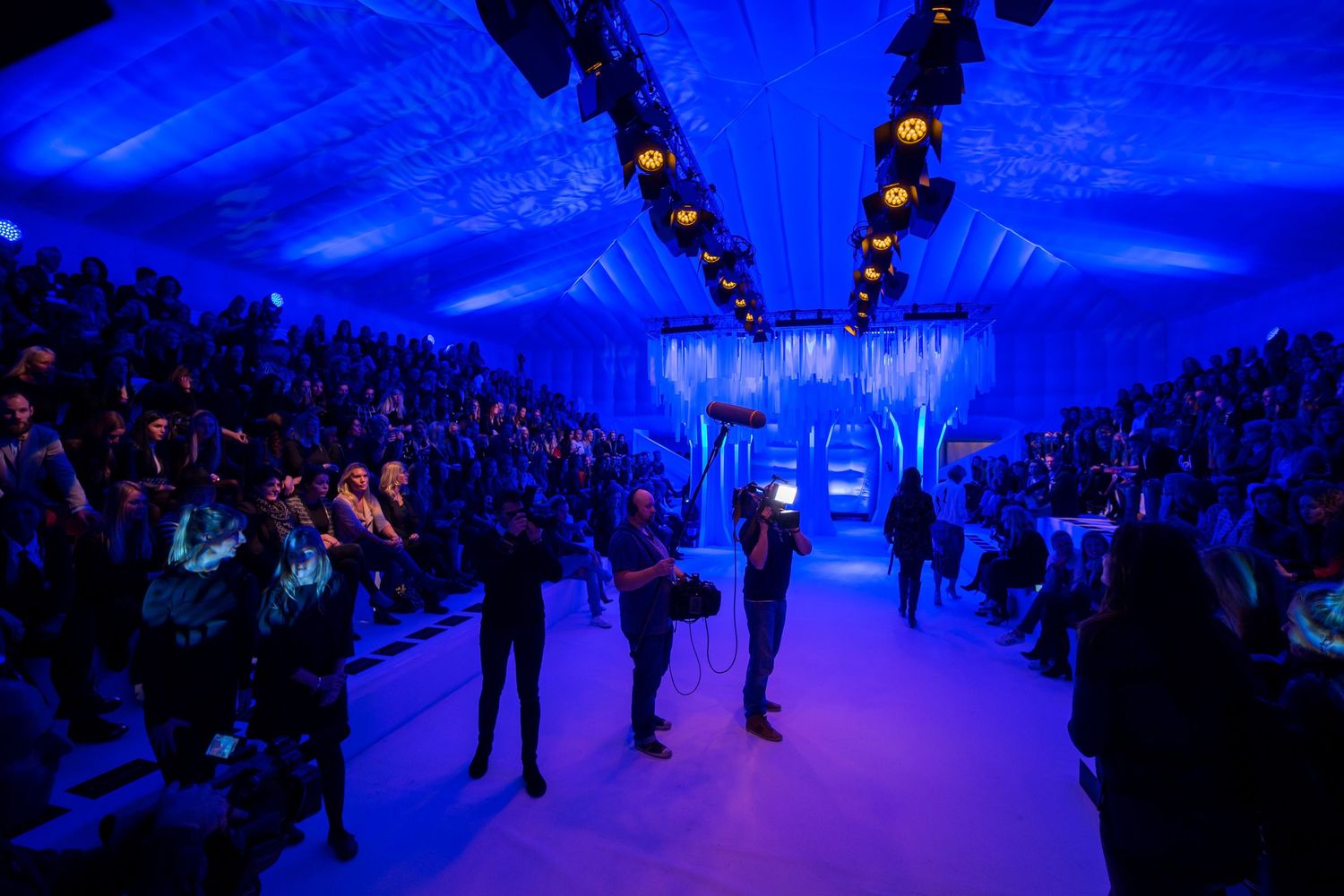 A fashion show runway bathed in blue light with a large audience seated on both sides.