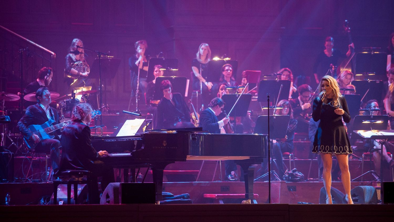 Female vocalist performs on stage with grand piano and full orchestra under purple stage lighting.