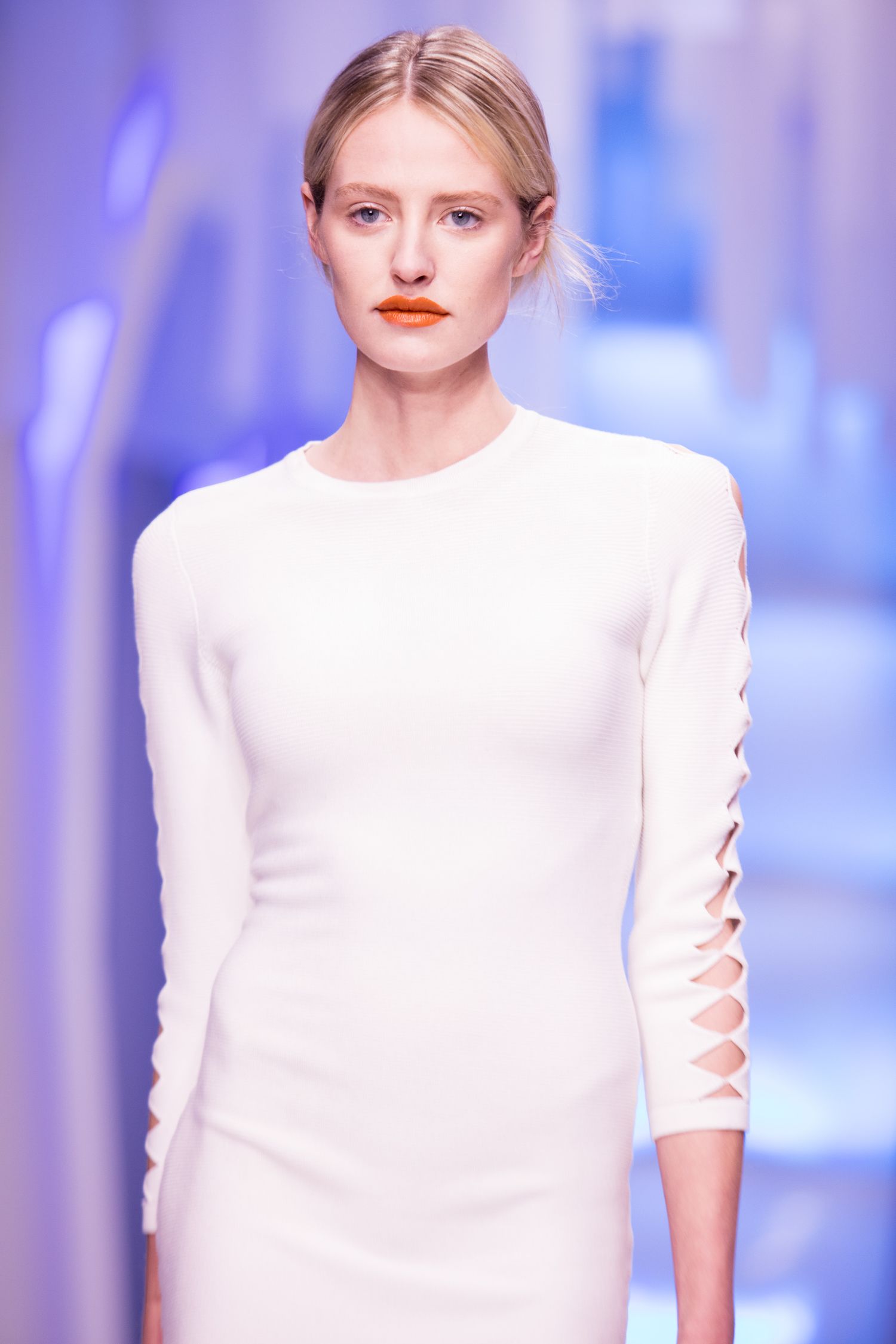 Model walks runway in white long-sleeve dress with cutout detail sleeves and bold orange lips.