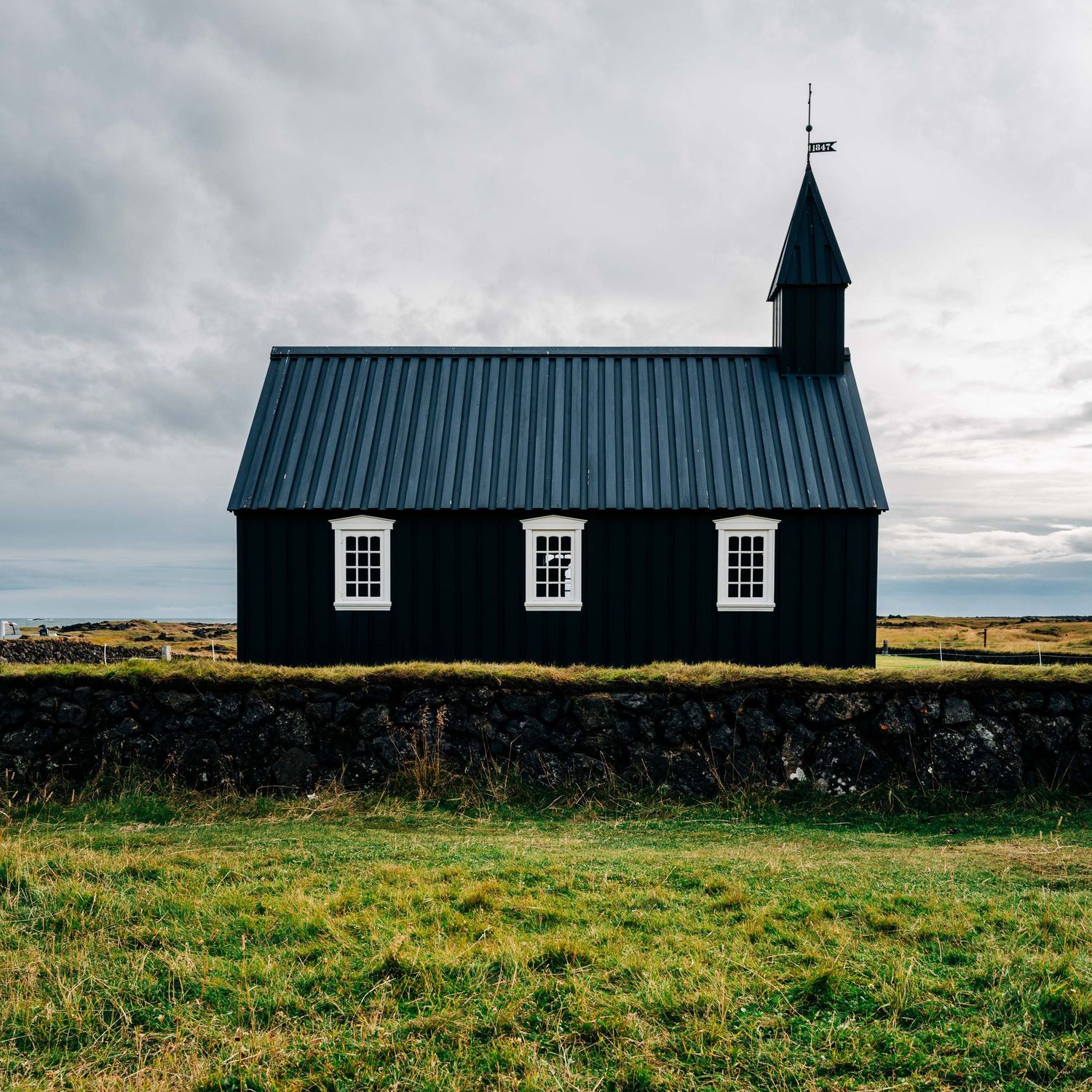 A black wooden church with white windows stands isolated against a moody sky in the Icelandic countryside.