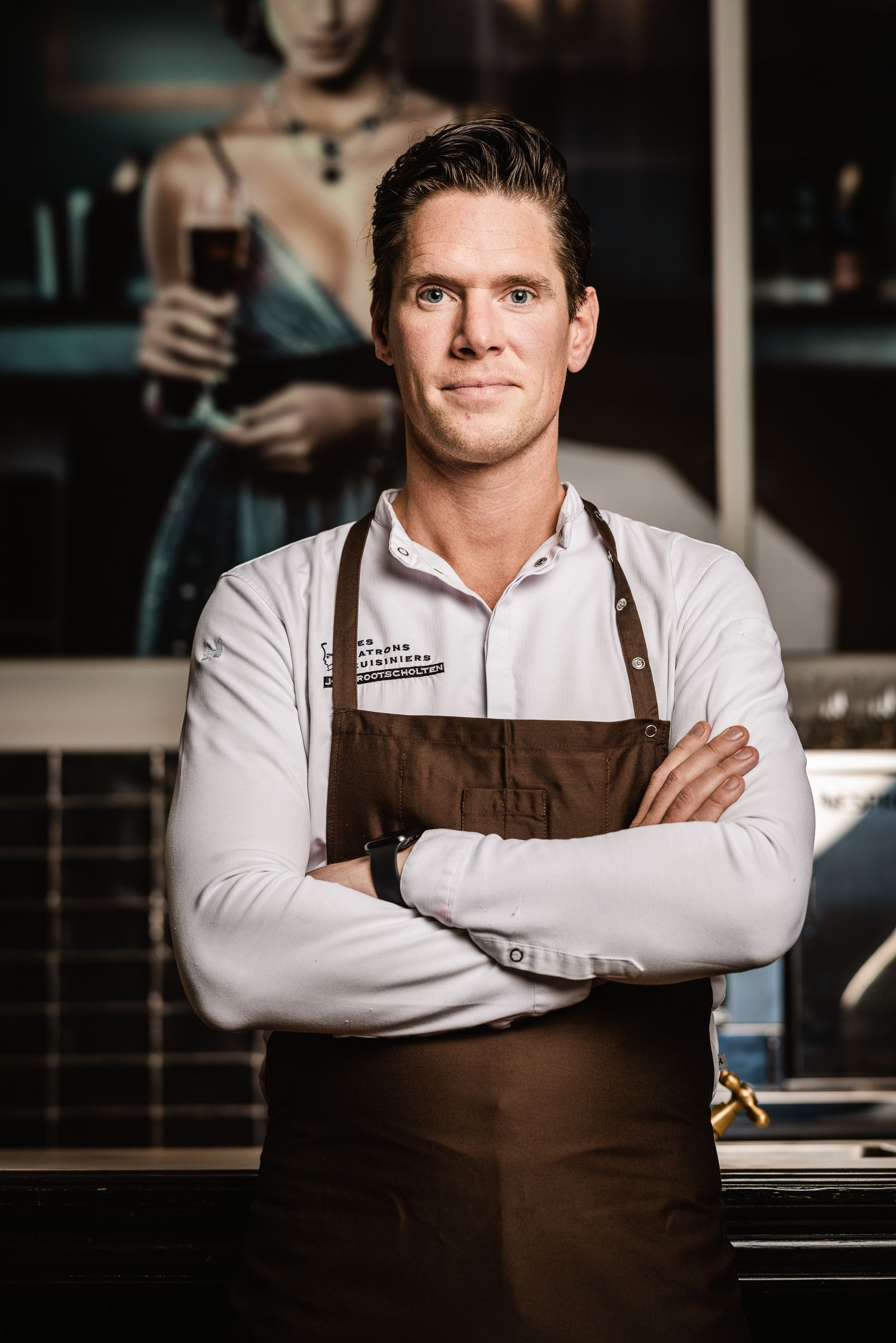 Restaurant worker in brown apron and white shirt poses with crossed arms in front of bar counter.