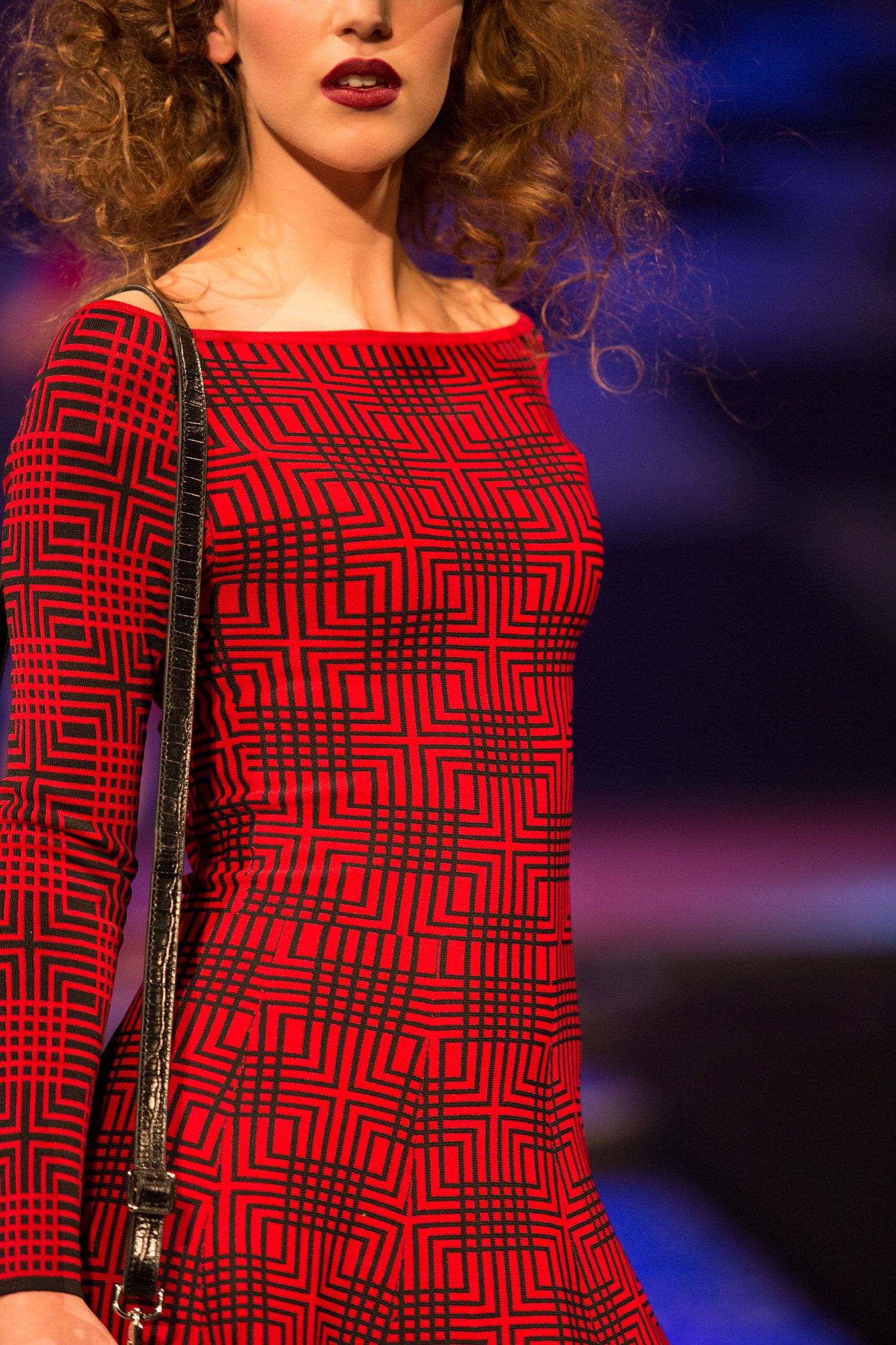 Model wearing a red and black geometric patterned dress on a fashion runway, carrying a black handbag.