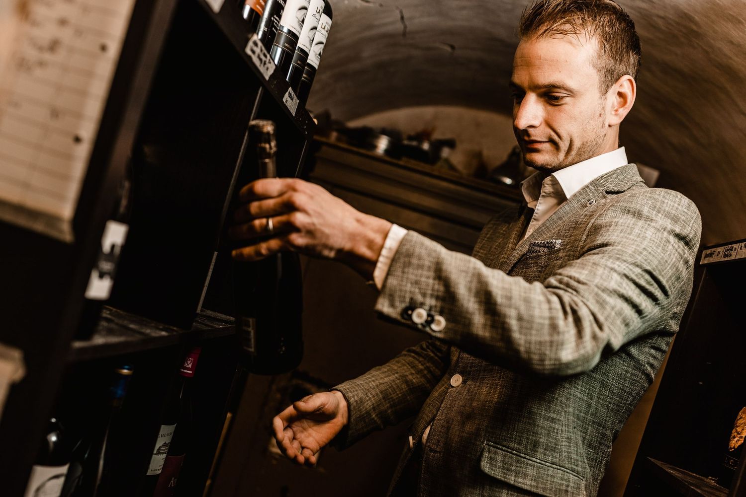 A person in a gray suit pours a cocktail into a glass at a dimly lit bar.