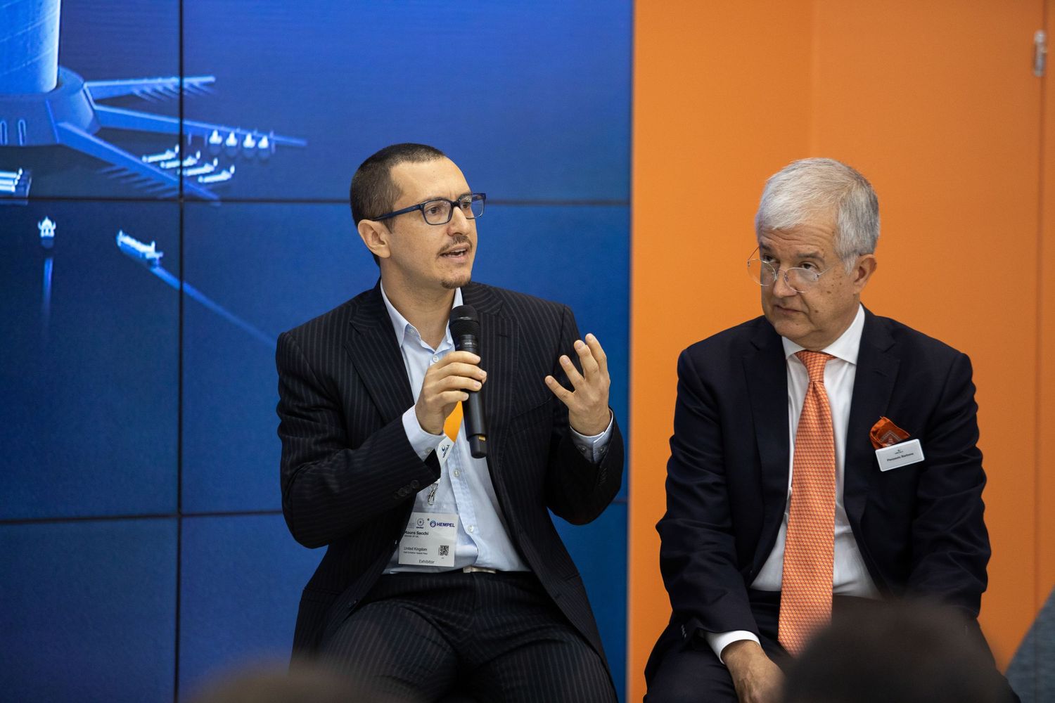 Business professionals participate in a panel discussion against blue and orange backdrop during a conference event.