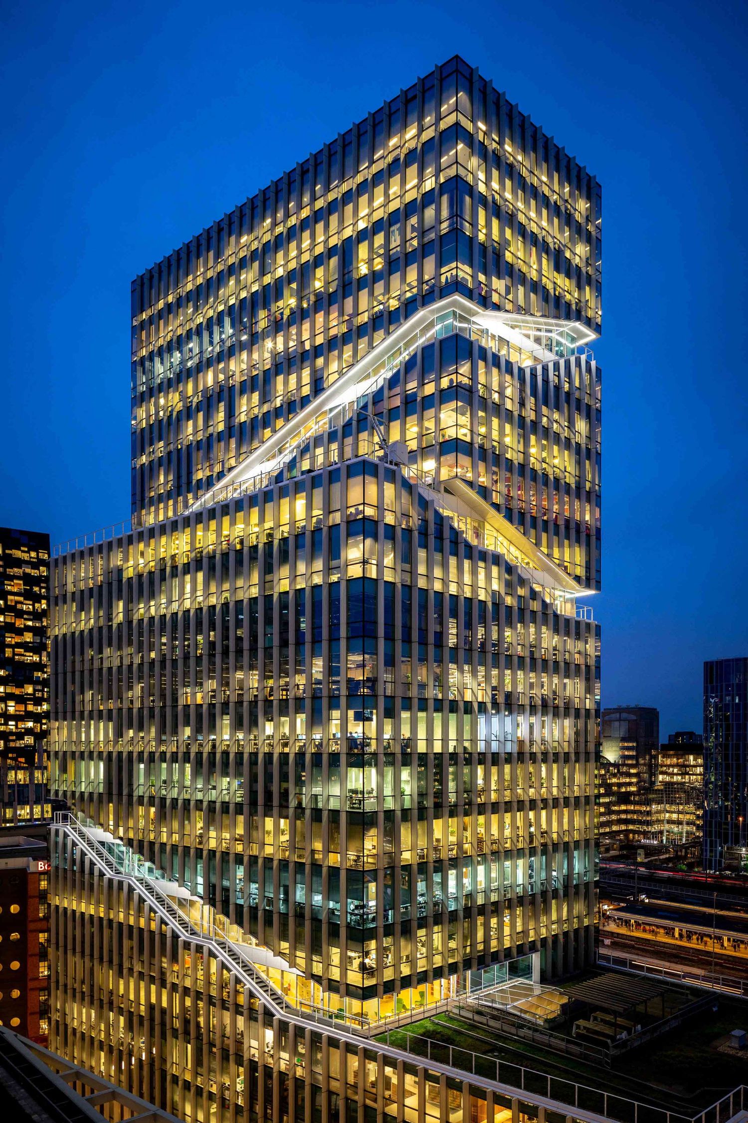 Modern office building with illuminated zigzag pattern facade glows against deep blue twilight sky in urban setting.