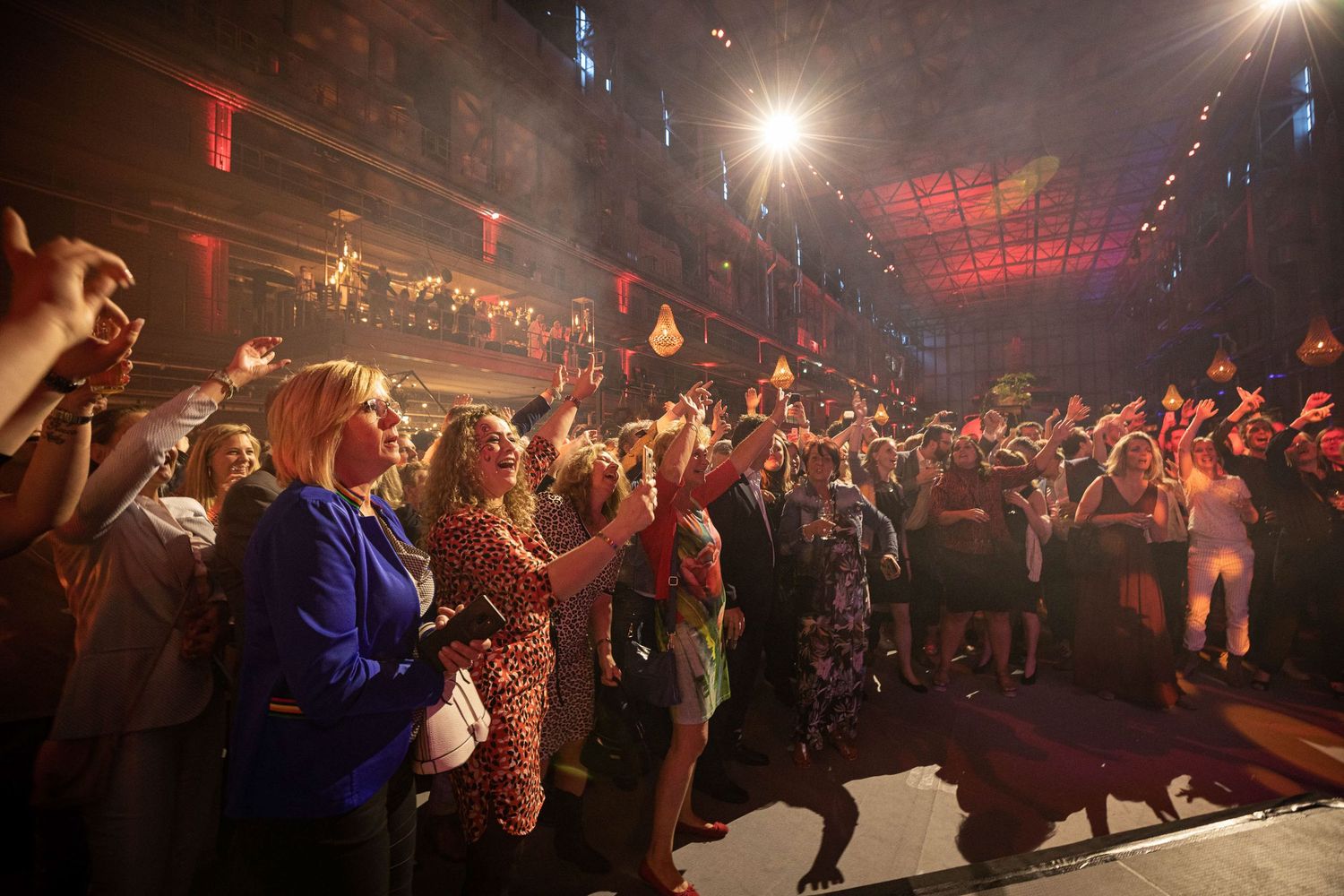A lively concert crowd dances and celebrates in a dimly lit venue with dramatic stage lighting.