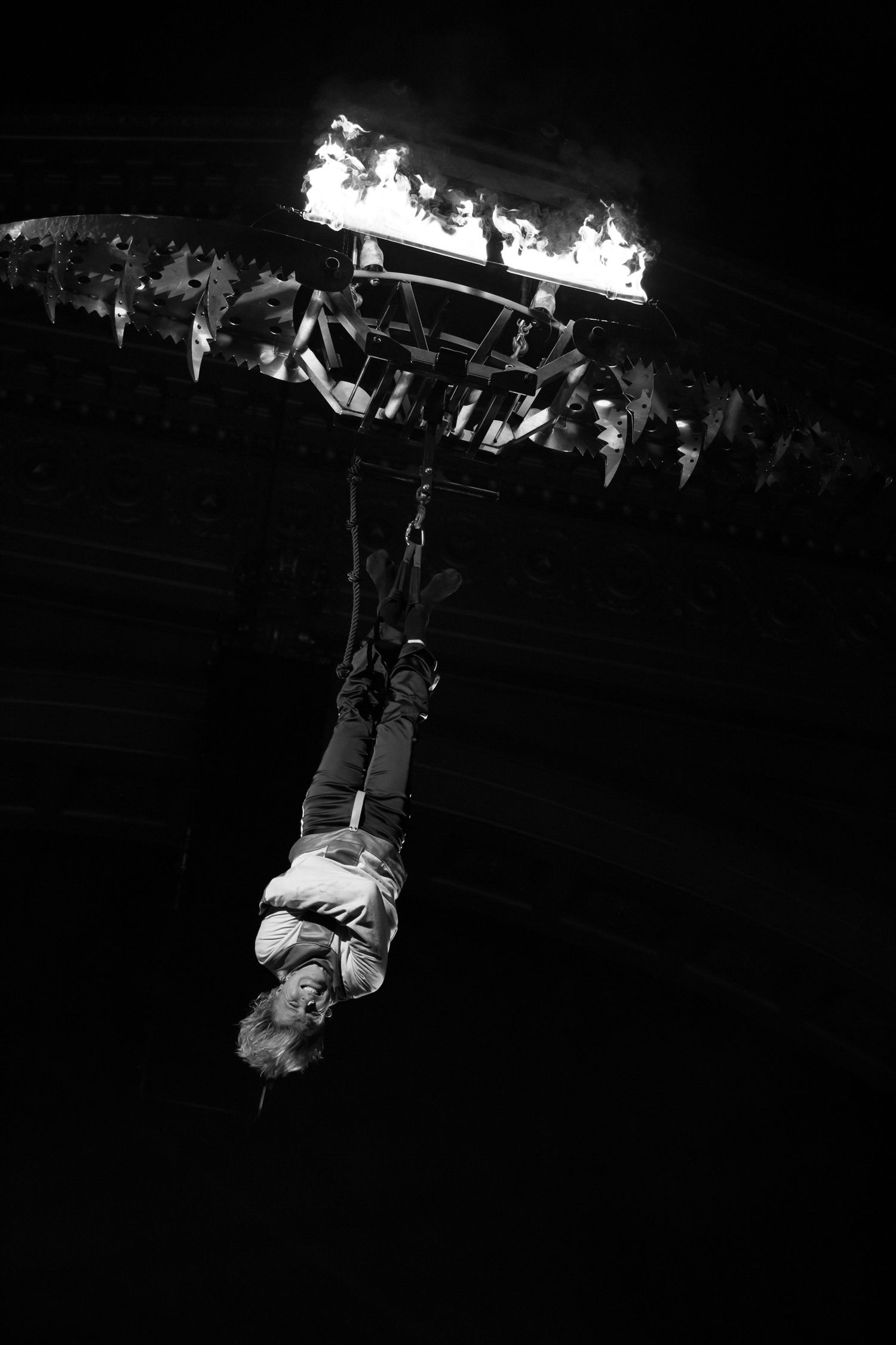 Black and white photo of a person hanging upside down from a flaming structure during a dramatic performance.