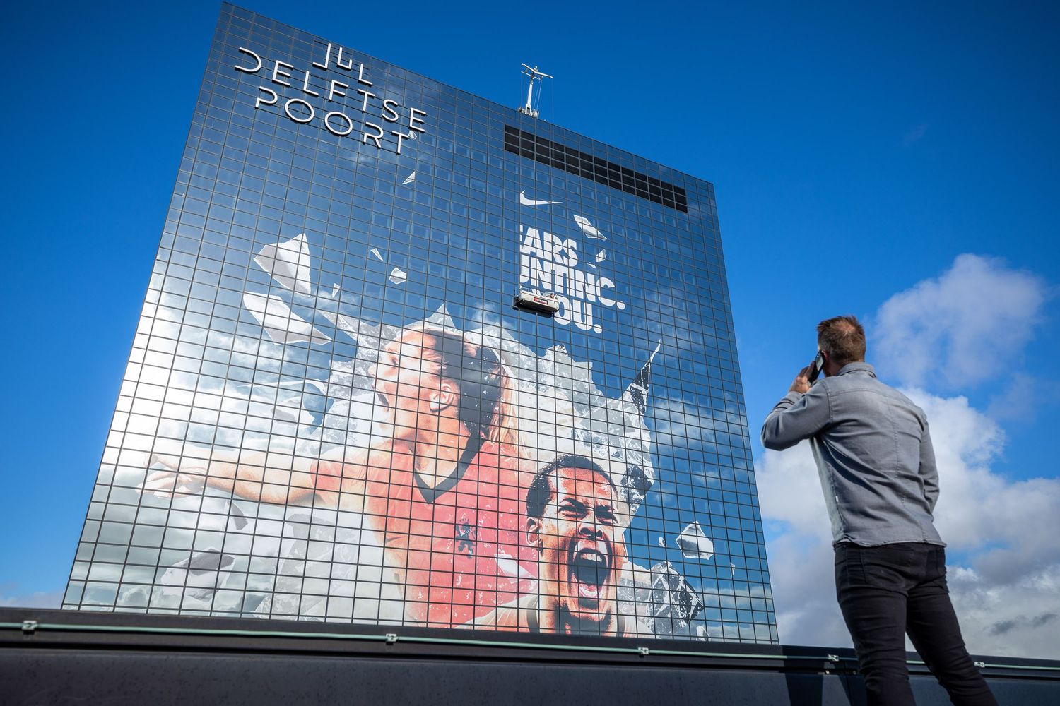 A man photographs a large Nike advertisement mural on a glass building under a blue sky.