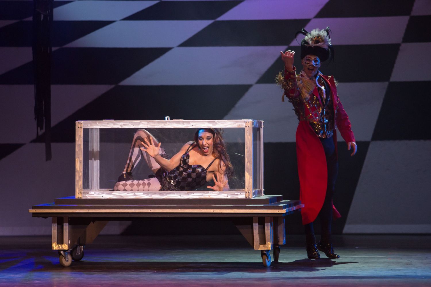 Magician in red coat performs box illusion trick with assistant on stage with checkered backdrop.