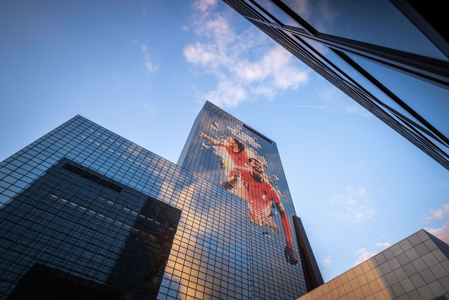 Towering glass skyscraper with a large red artistic mural reflecting blue sky and clouds in an urban cityscape.