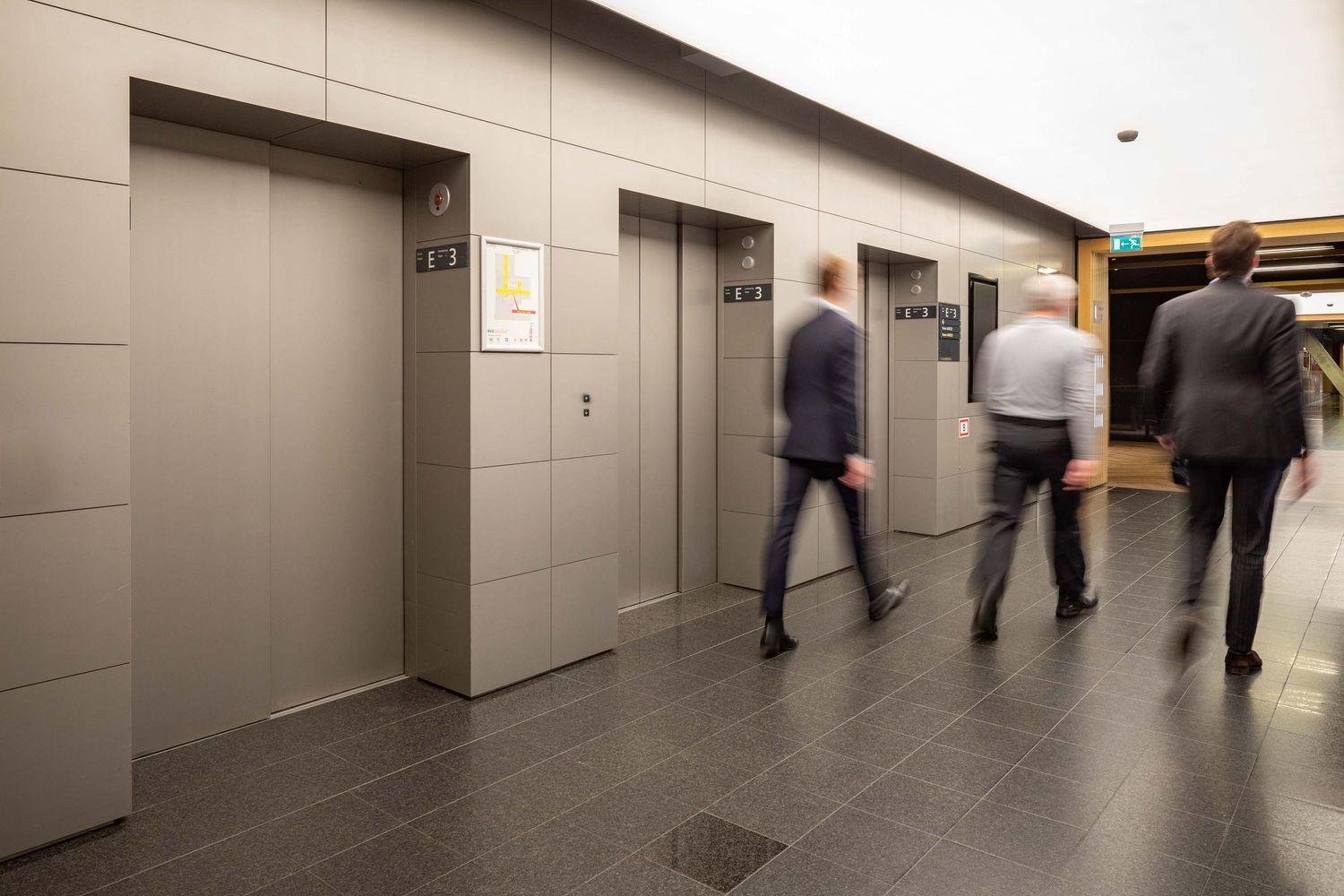 Business professionals walk through a modern office hallway with gray walls and bright lighting.