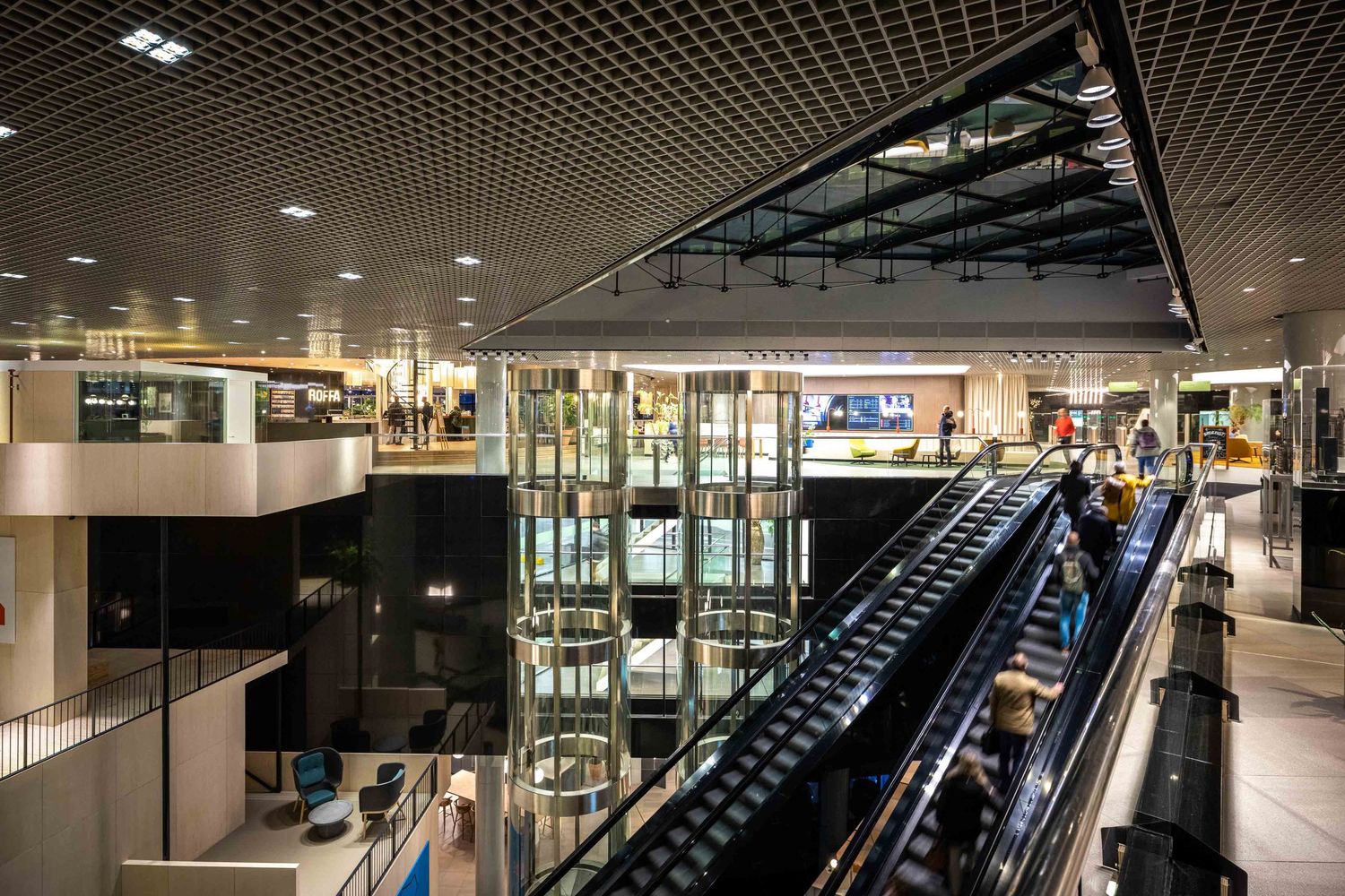 Modern shopping mall interior with escalators, glass railings, and open atrium design under grid pattern ceiling lighting.