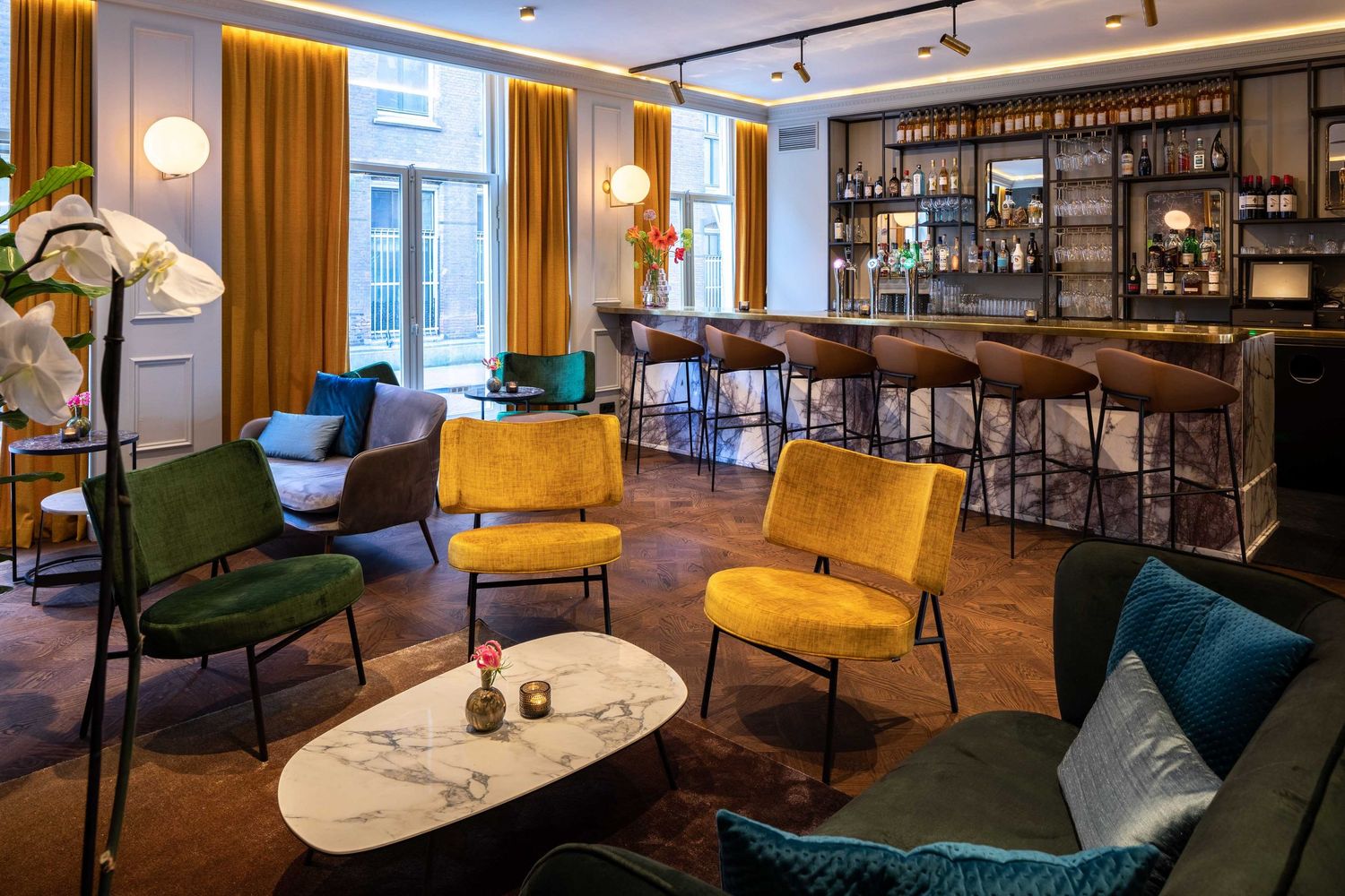 Stylish hotel lounge with velvet armchairs in jewel tones, marble tables, and an elegant bar with warm lighting and gold accents.