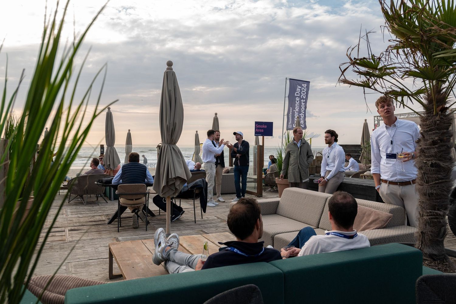 Outdoor event space with palm trees and people gathering near the waterfront at sunset.