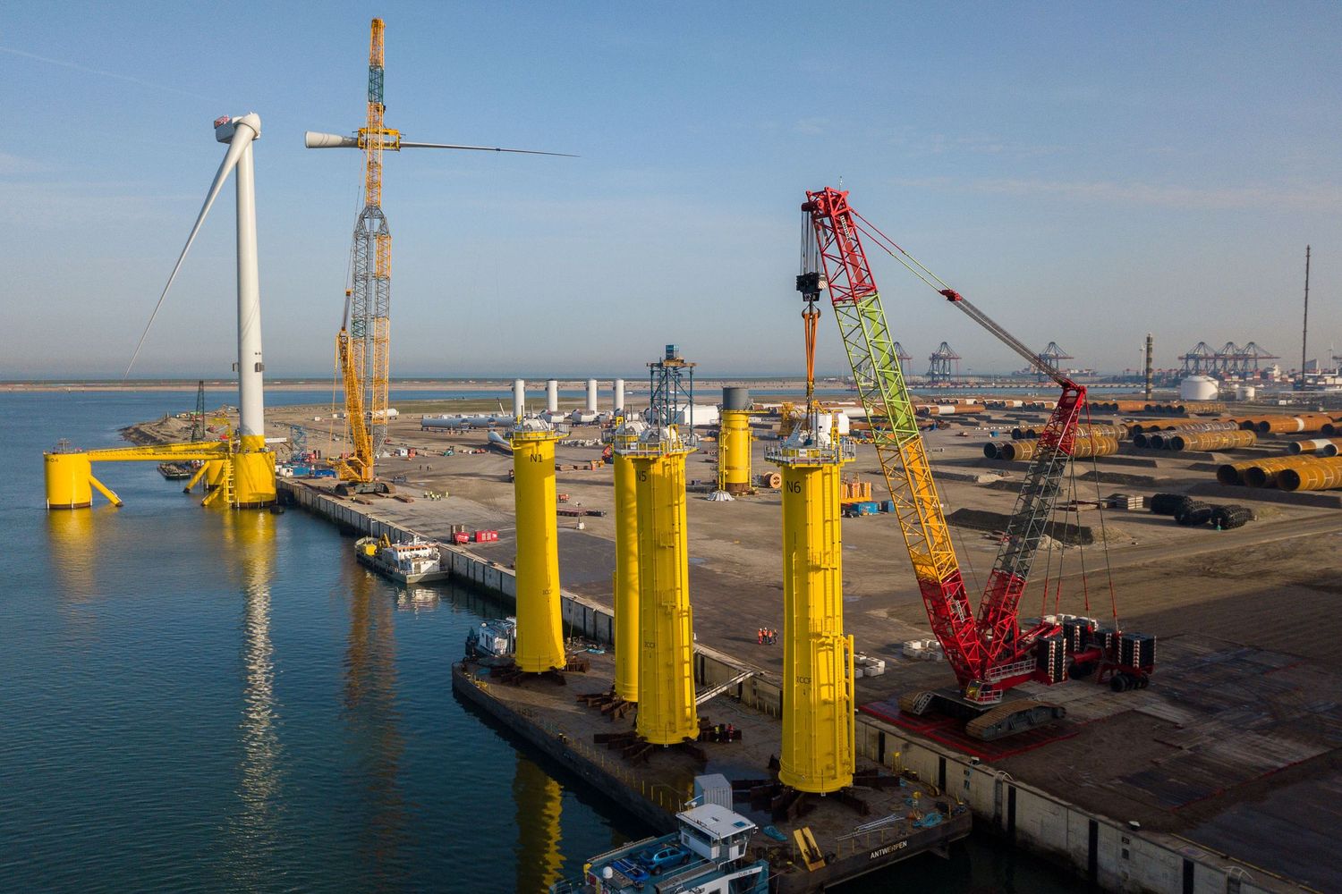 Offshore wind turbine installation at industrial port with yellow foundations and cranes under clear sky.