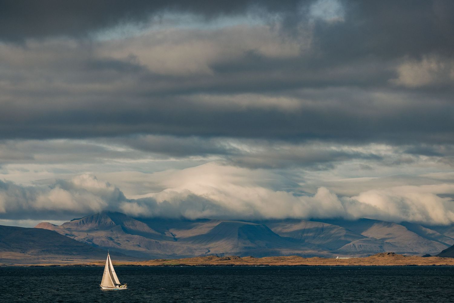 Dramatic stormy clouds roll over rugged mountains and coastal waters as a small sailboat glides across the horizon.