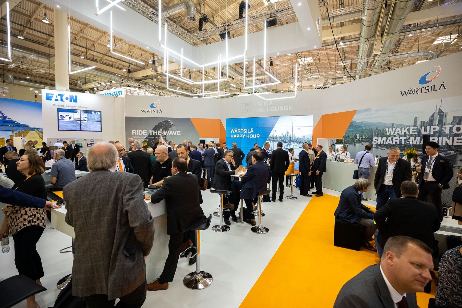 Business professionals gather at a crowded trade show exhibition with bright orange and blue corporate displays.