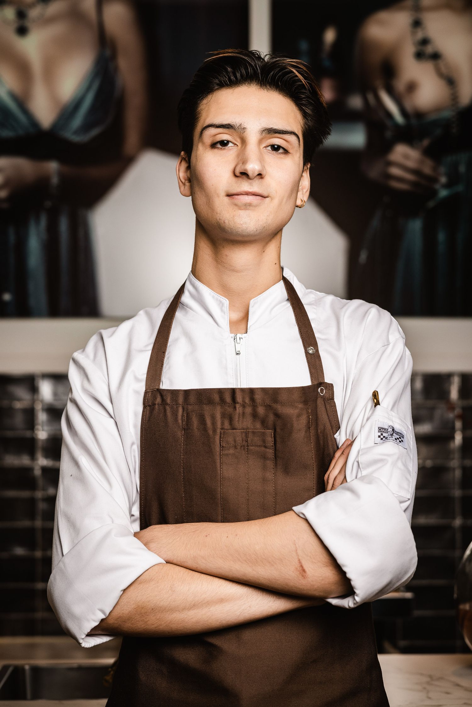 Professional chef in white coat and brown apron stands confidently with crossed arms in restaurant kitchen.