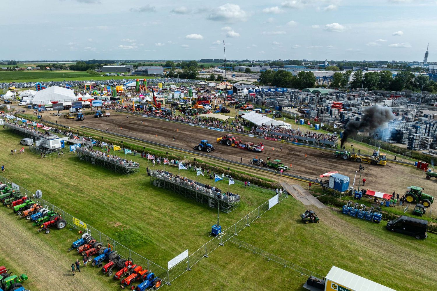 Aerial view of a tractor pulling competition at a rural fairground with crowds, colorful tractors, and smoke on a cloudy day.