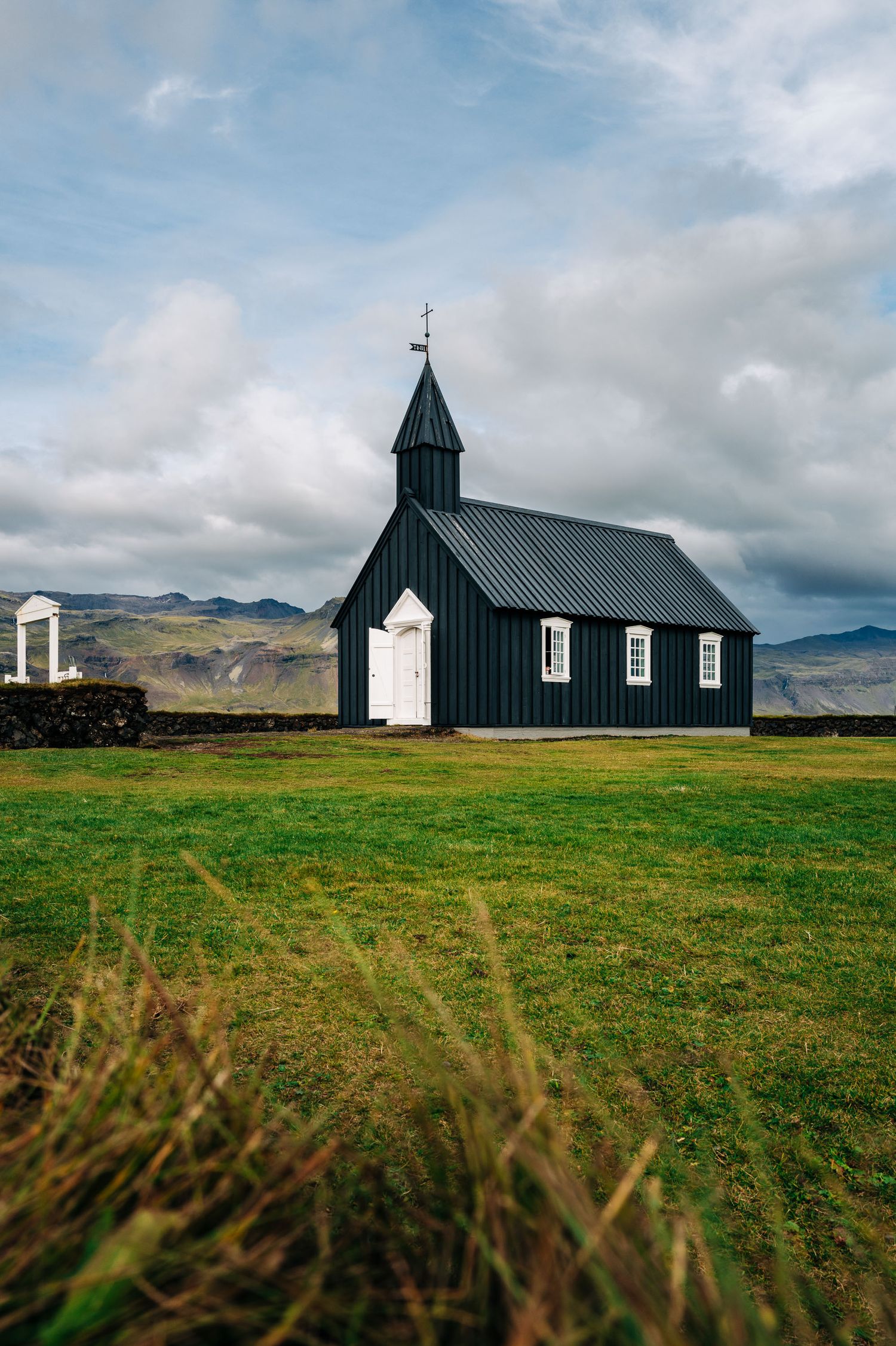 A black wooden church with a steeple stands solitary on a green field against a cloudy mountain landscape in Iceland.