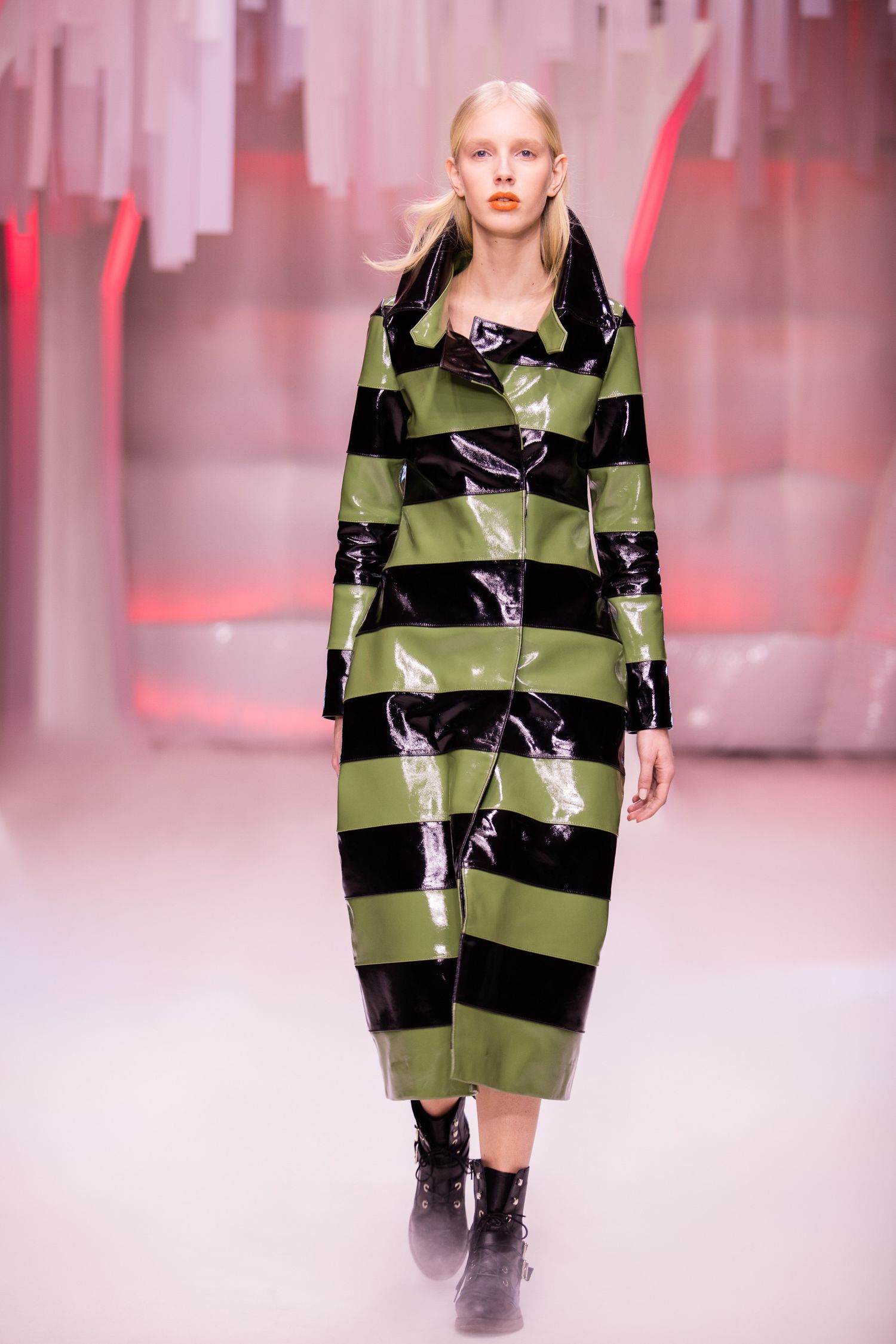 Model walks runway in glossy green and black horizontal striped vinyl coat with black ankle boots at fashion show.