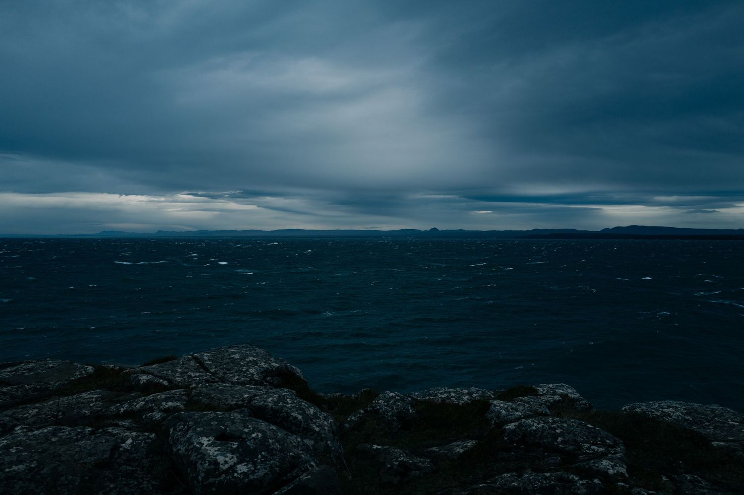 Dark moody seascape with rocky coastline and stormy clouds over deep blue waters at dusk.