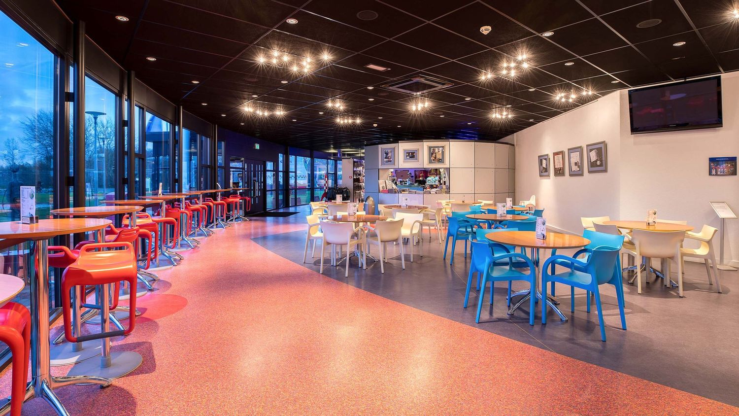 Modern cafeteria with colorful chairs, large windows, pendant lights, and an open dining area with tables on a red terrazzo floor.
