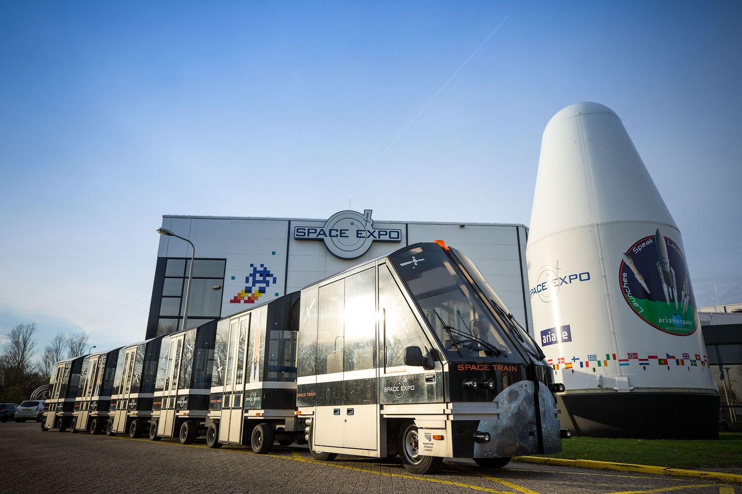 Space Expo museum exterior with rocket replica and transport vehicles under a clear blue sky.
