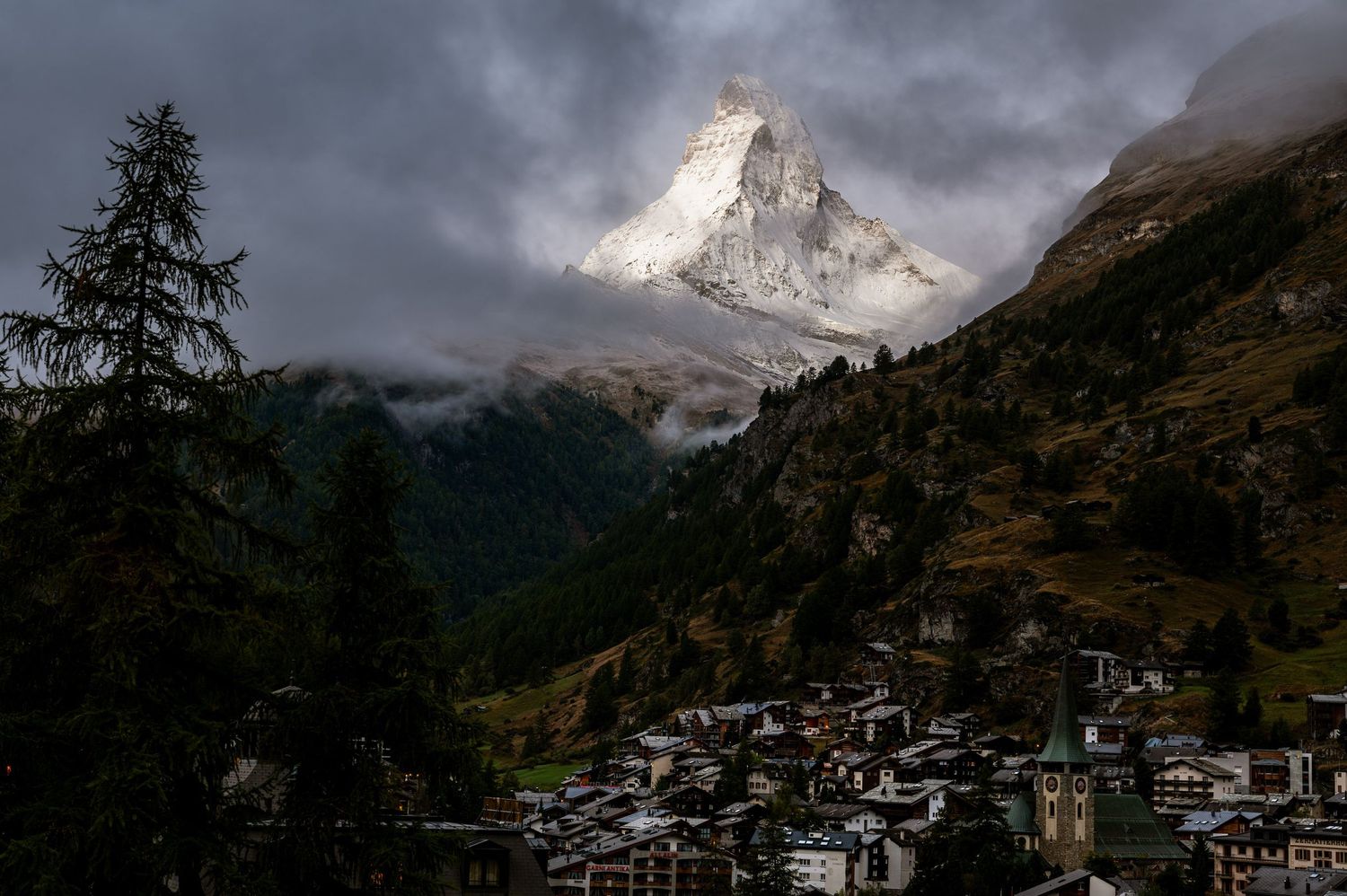 Snow-capped Matterhorn peak towers over alpine village under stormy skies.