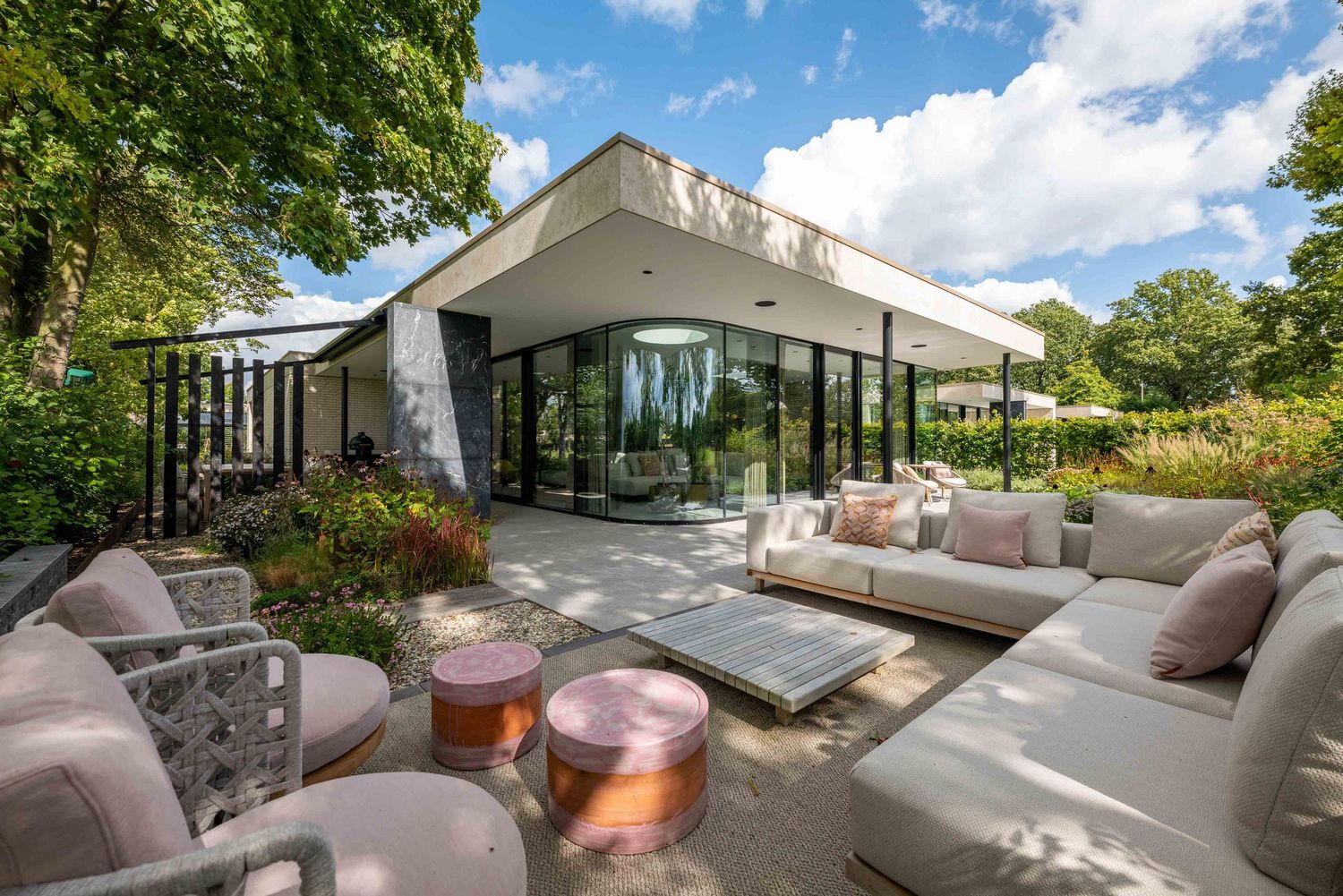 Modern outdoor patio with comfortable seating and glass railings overlooking a lush garden landscape on a sunny day.