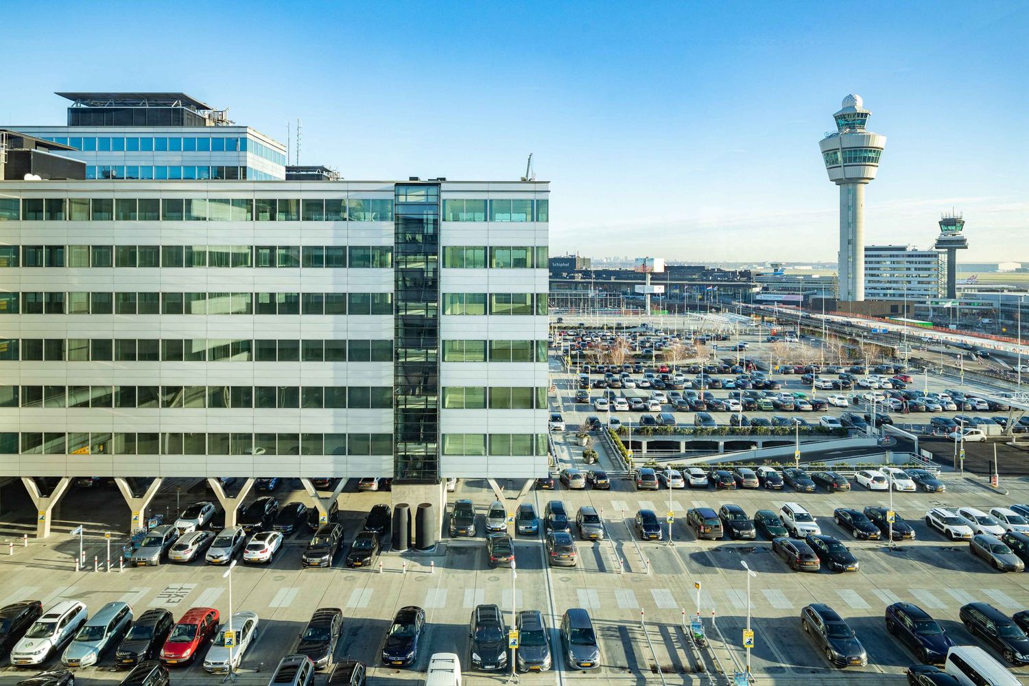 Airport terminal exterior with control tower and rows of parked cars in parking lot.