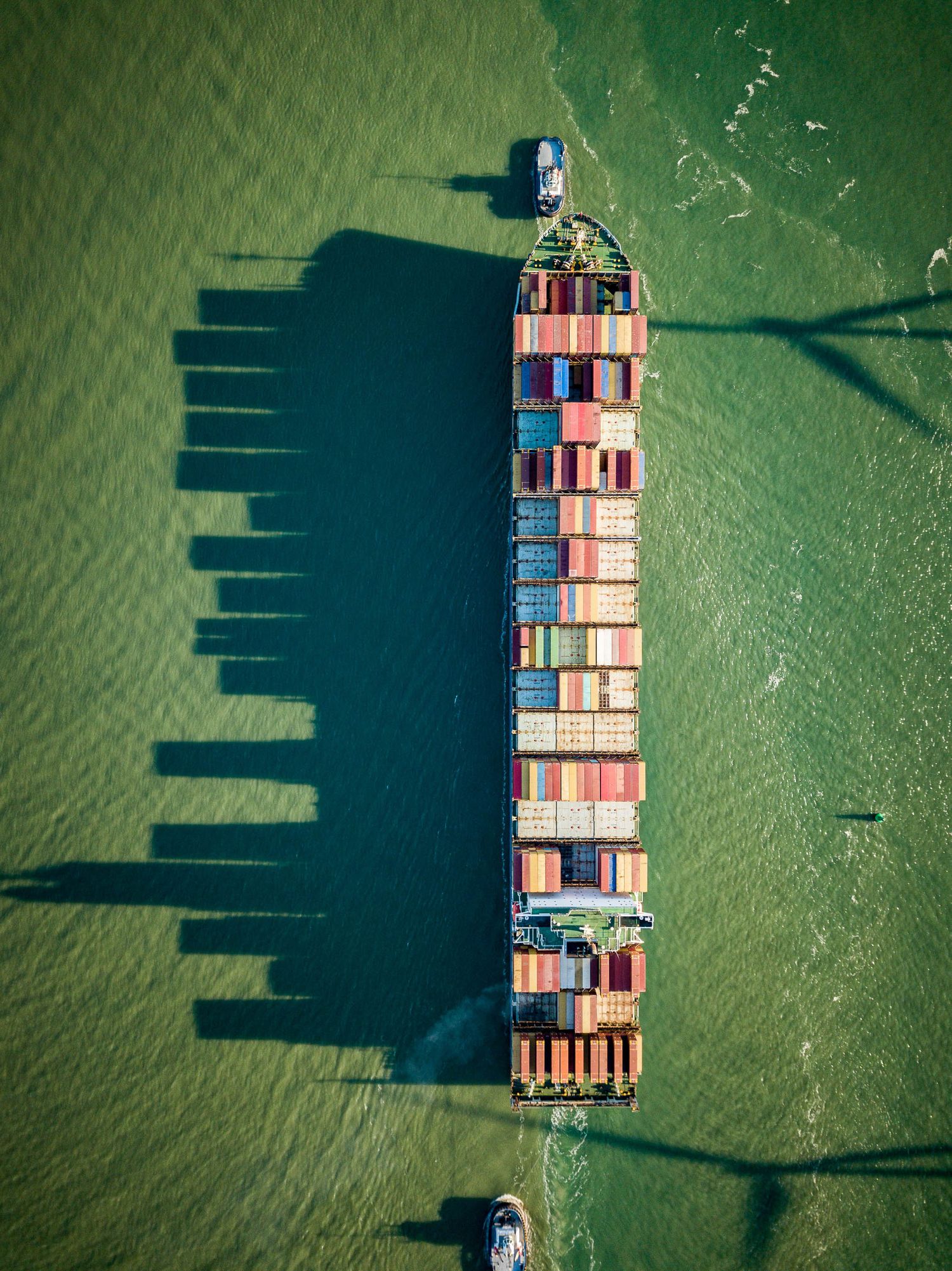 Aerial view of a large cargo container ship loaded with colorful shipping containers casting shadows on green ocean water.