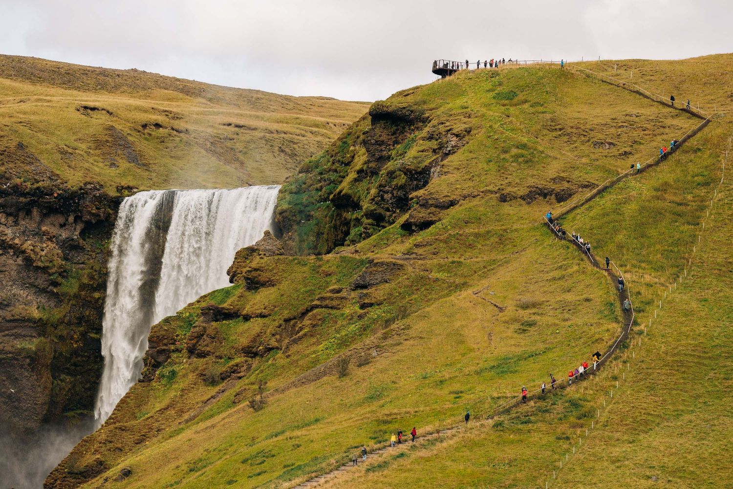 Hikers walking along a scenic mountain trail beside a cascading waterfall in green rolling hills.