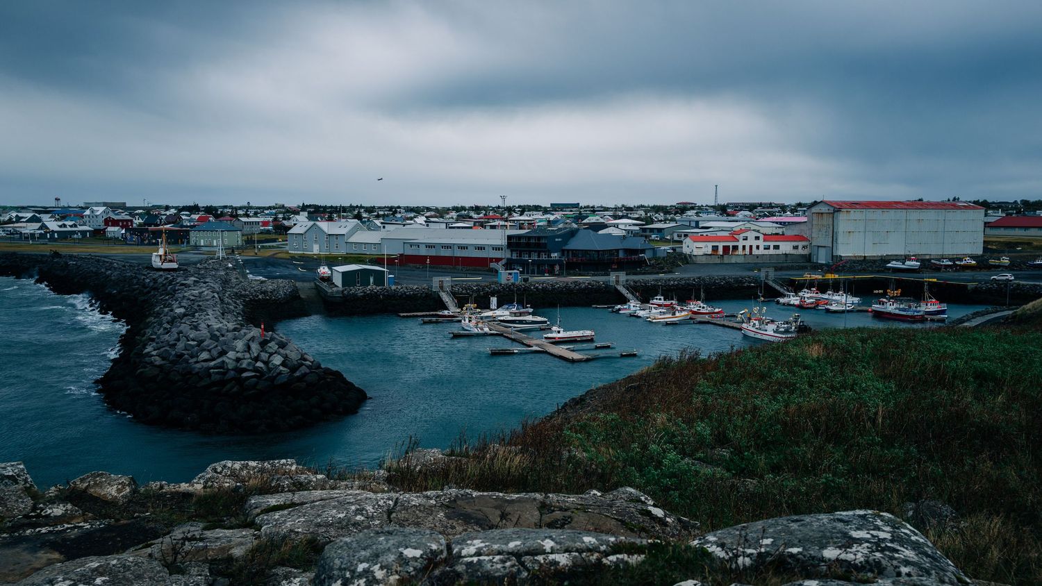 Scenic harbor view of a coastal town with boats docked in a small bay surrounded by rocky shores and industrial buildings.