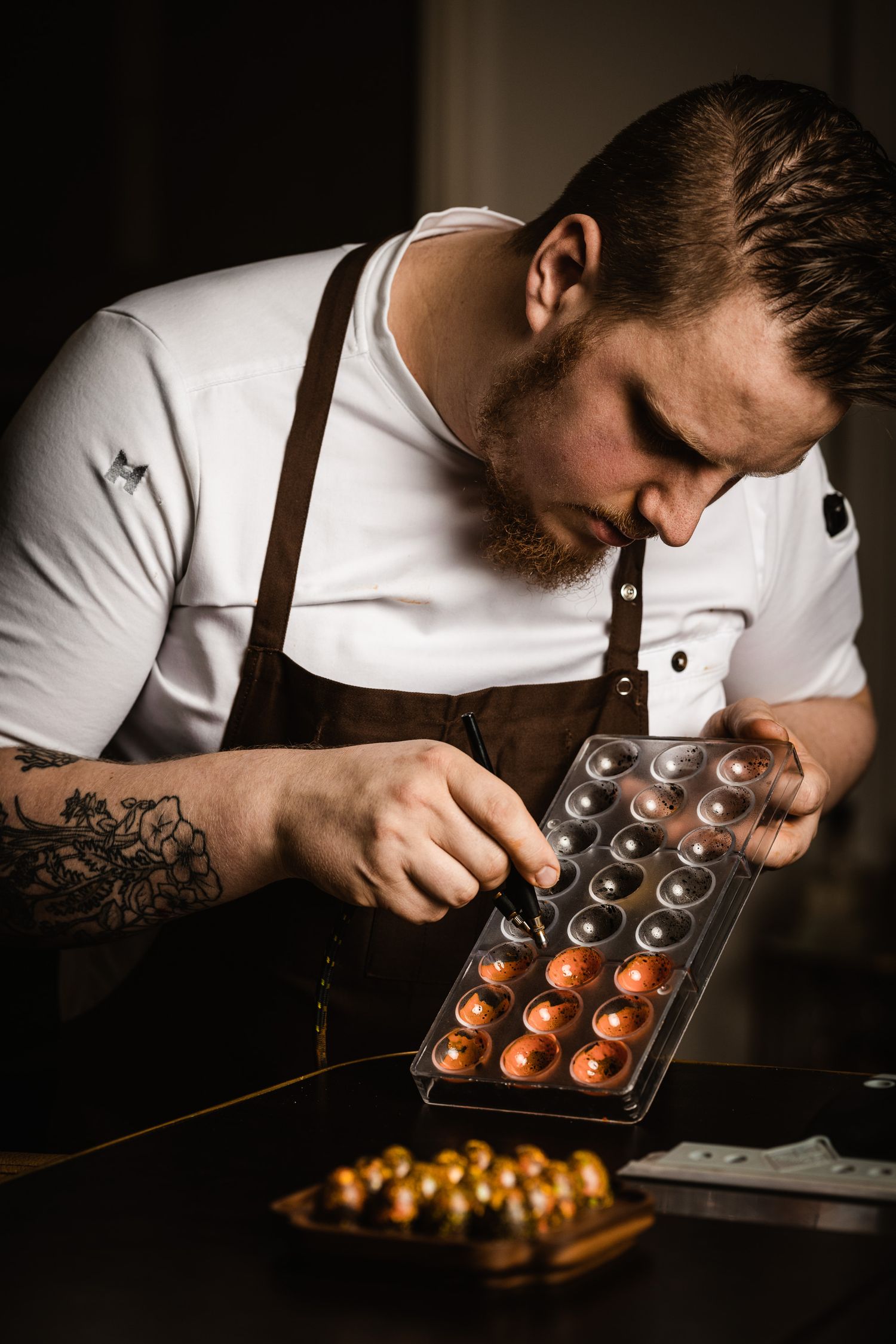 A chocolatier in a white shirt and leather apron carefully crafts artisanal chocolates in decorative molds.