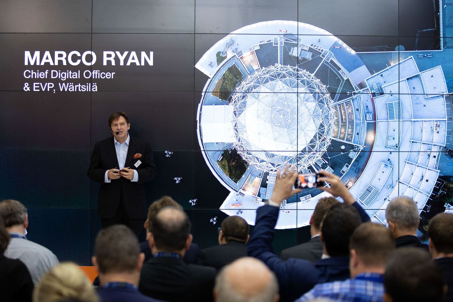 Speaker presents to a large audience in front of digital screens displaying data and analytics at a business conference.