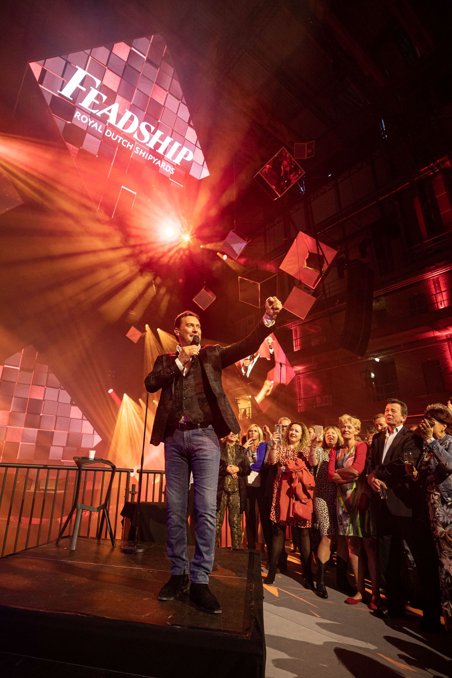 Performer on stage gestures enthusiastically under pink lighting at a live event with 'Leadership' signage and crowd visible.