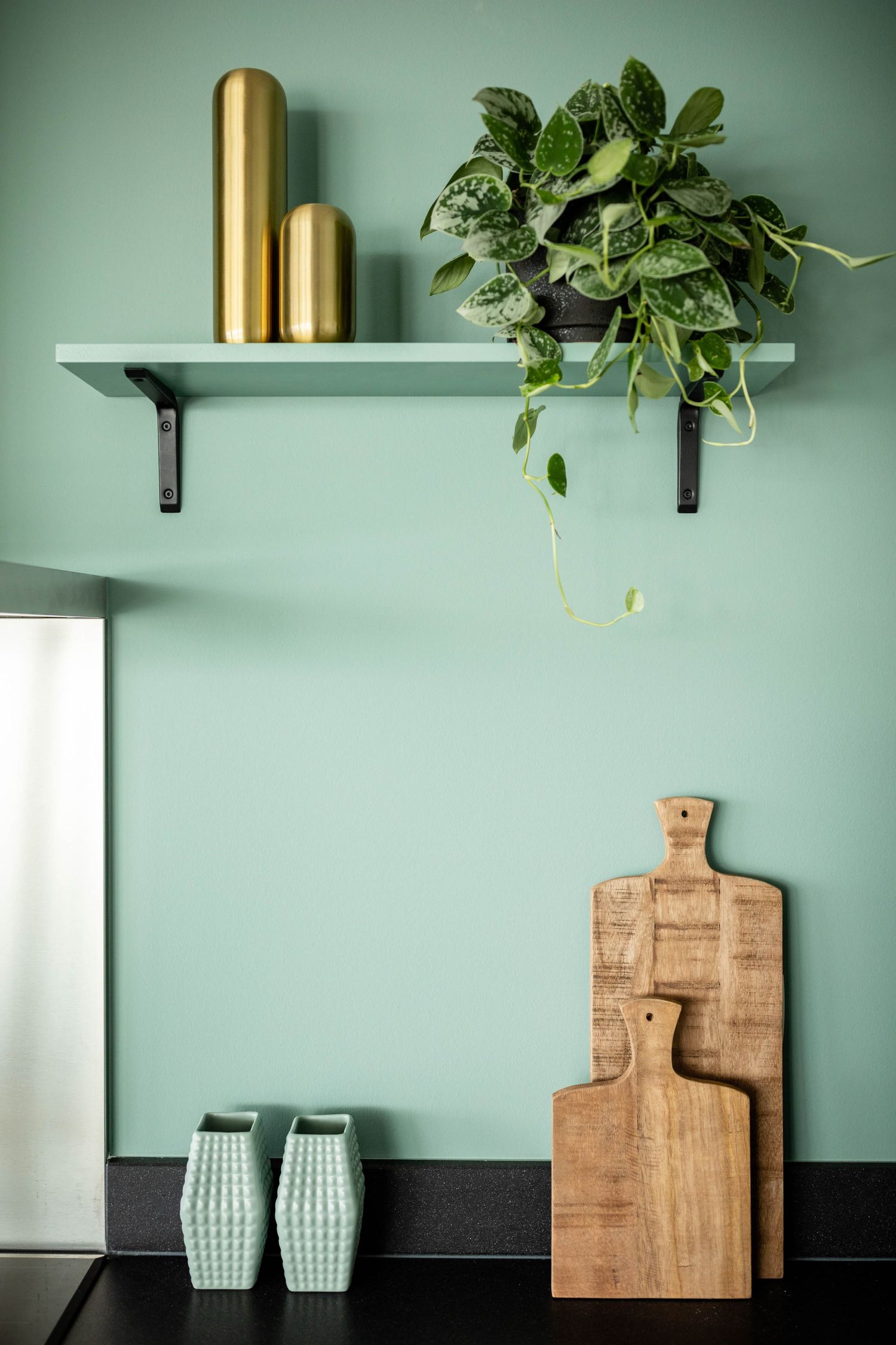 Minimalist kitchen decor featuring wooden cutting boards, green ceramic vases, and hanging plants against a sage green wall.