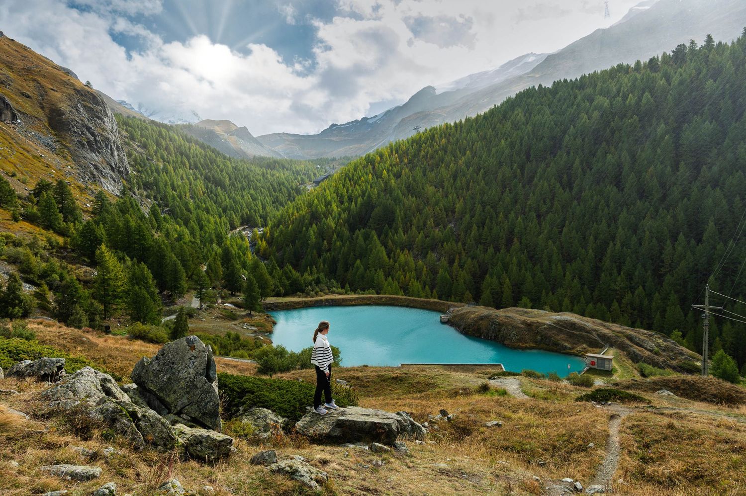 Panoramic mountain landscape with turquoise alpine lake surrounded by evergreen forests and rocky peaks.