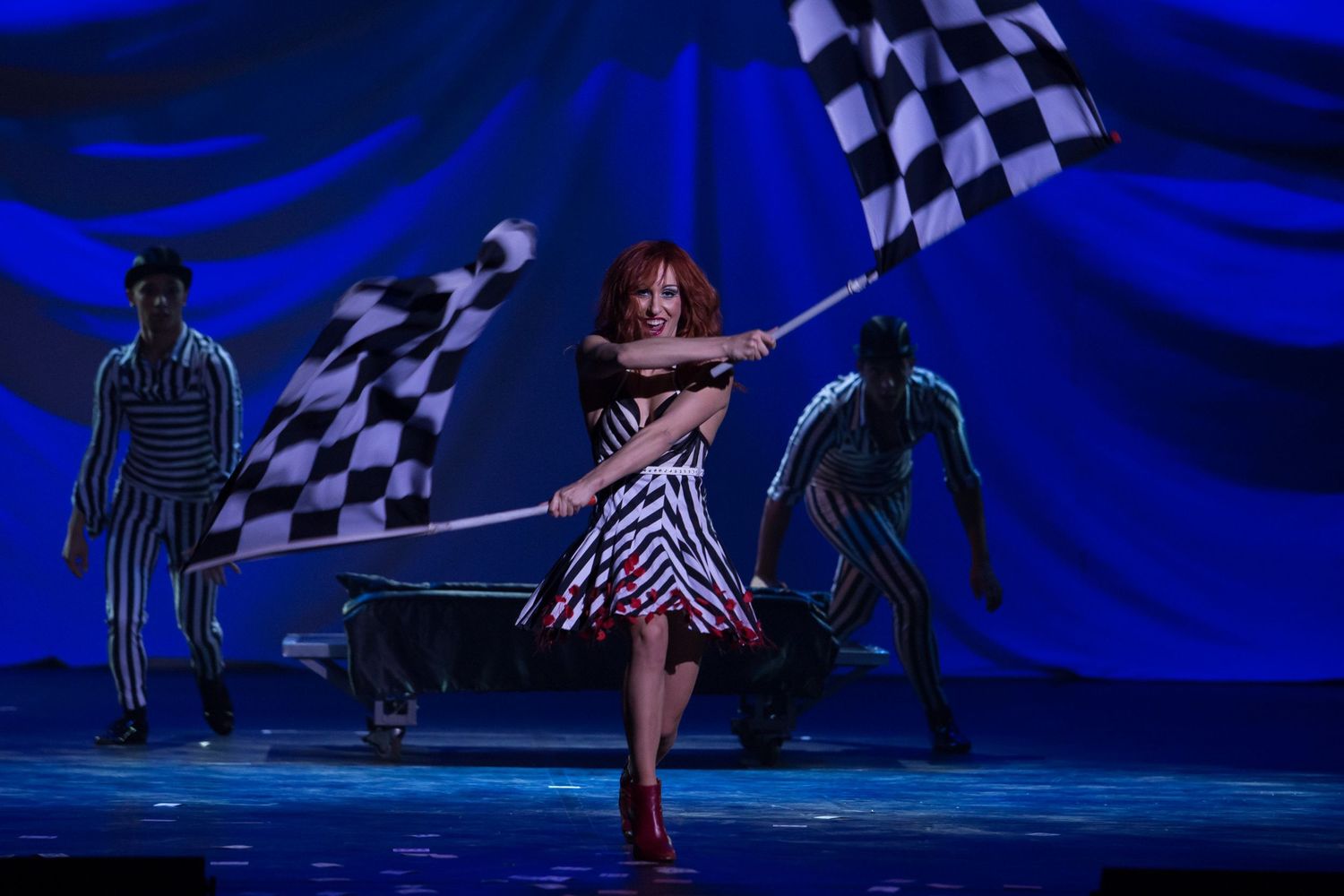 Performer in striped dress waves checkered flag on blue-lit stage with dancers in striped costumes behind her.