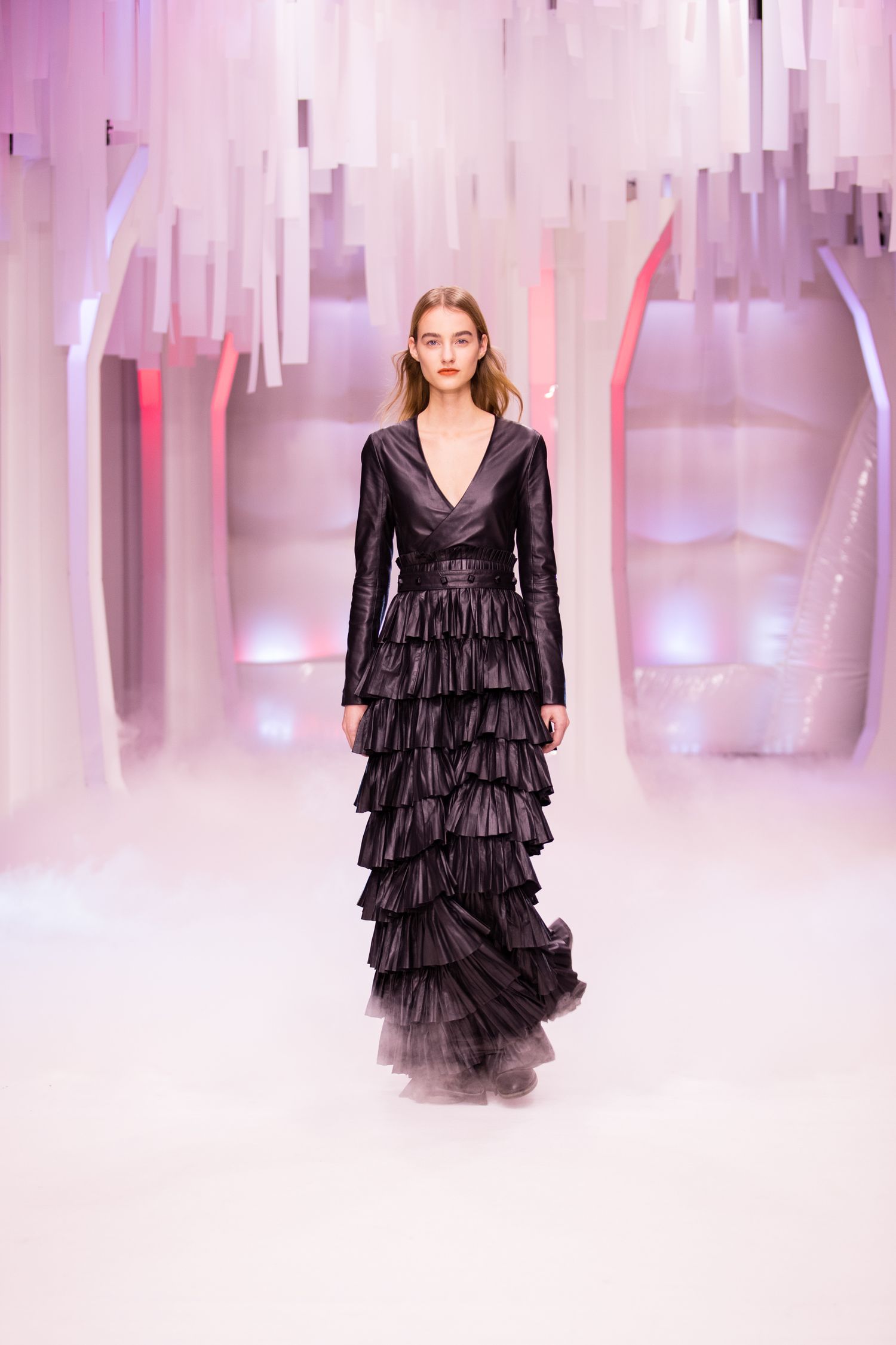 Model walks runway in black tiered ruffle gown with V-neck bodice, surrounded by pink misty light installation.