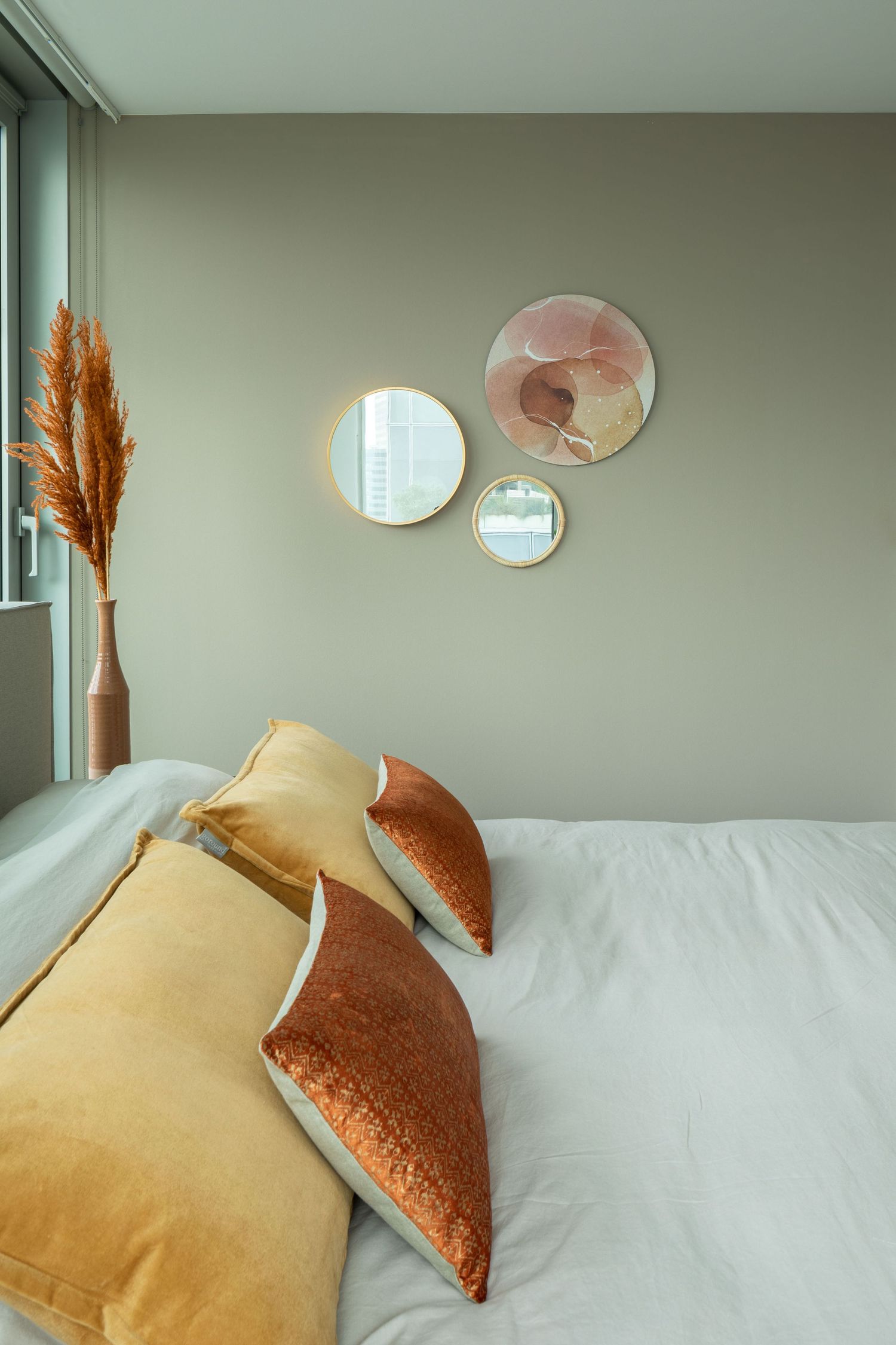 Modern bedroom interior with sage green walls, circular mirrors, pampas grass decor, and yellow and rust colored pillows.