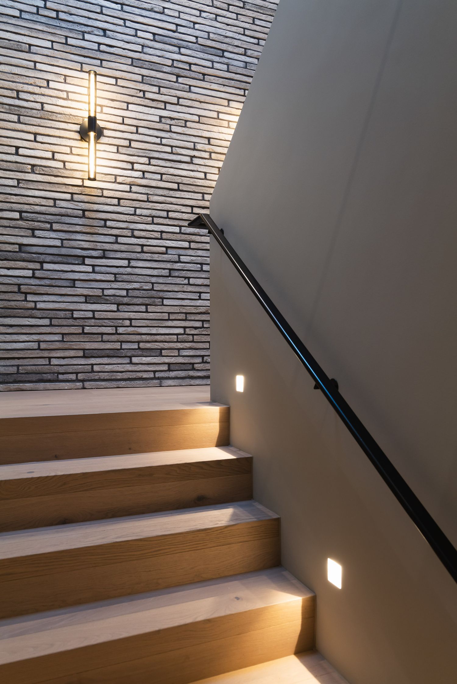 Modern wooden staircase with built-in LED lighting and black handrail against white brick wall creates elegant ambiance.