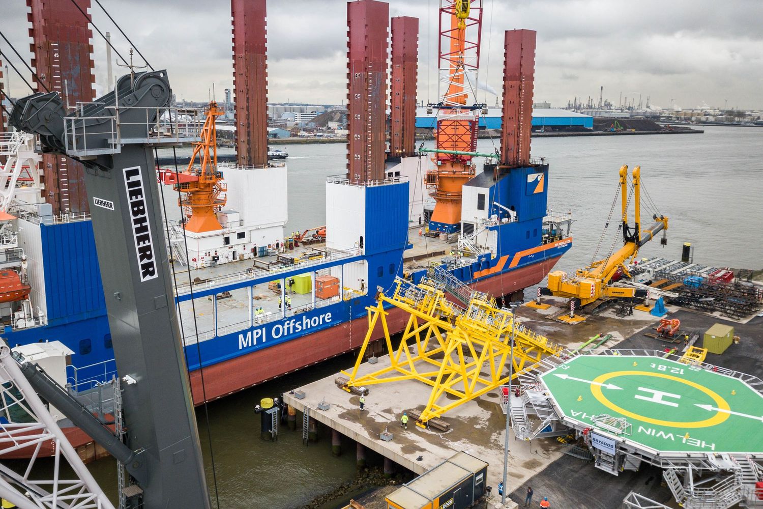 Industrial offshore vessel with cranes and helipad docked at a maritime facility.