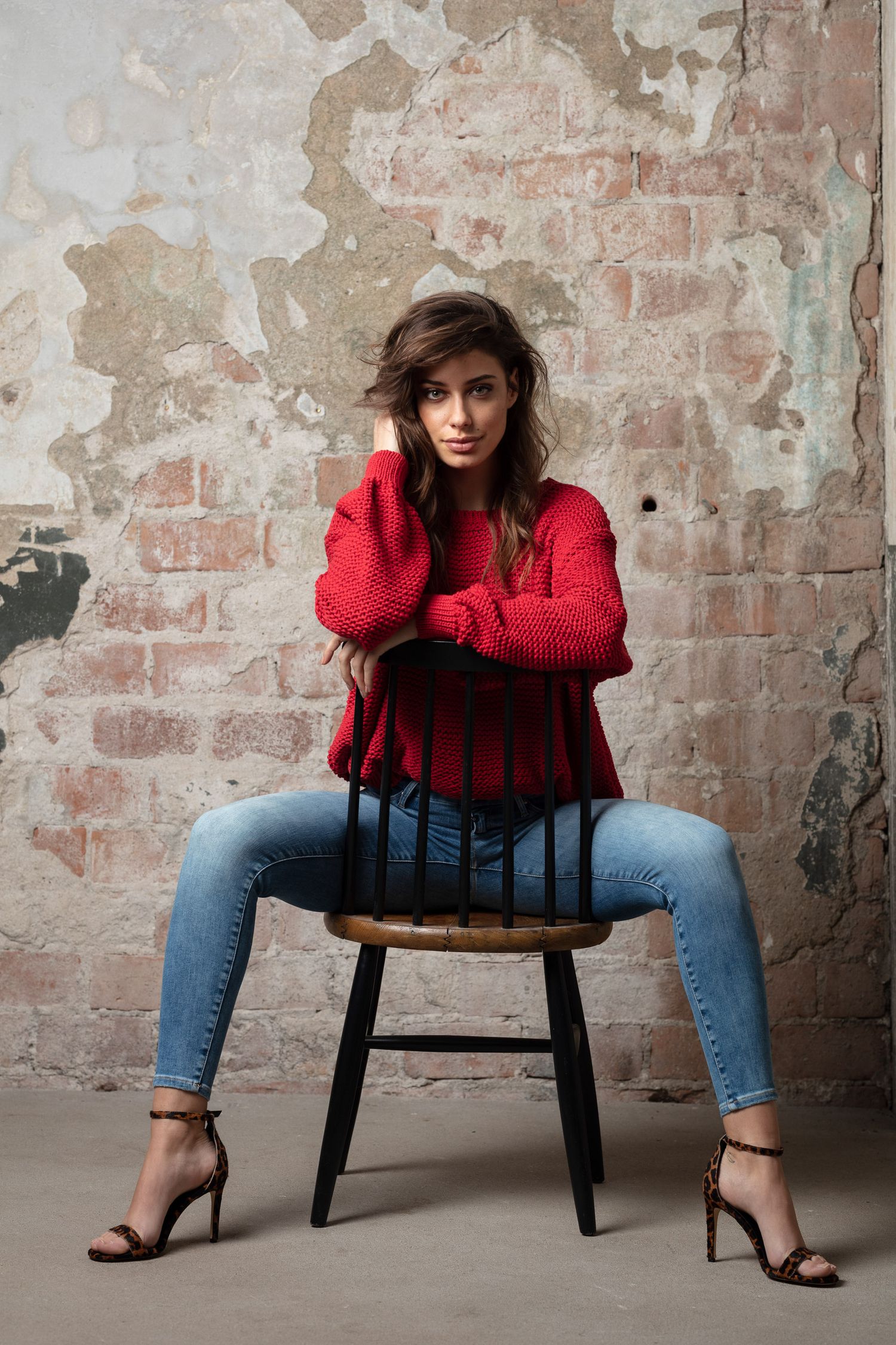 Fashion series featuring red sweater and jeans with wooden chair against rustic brick wall.
