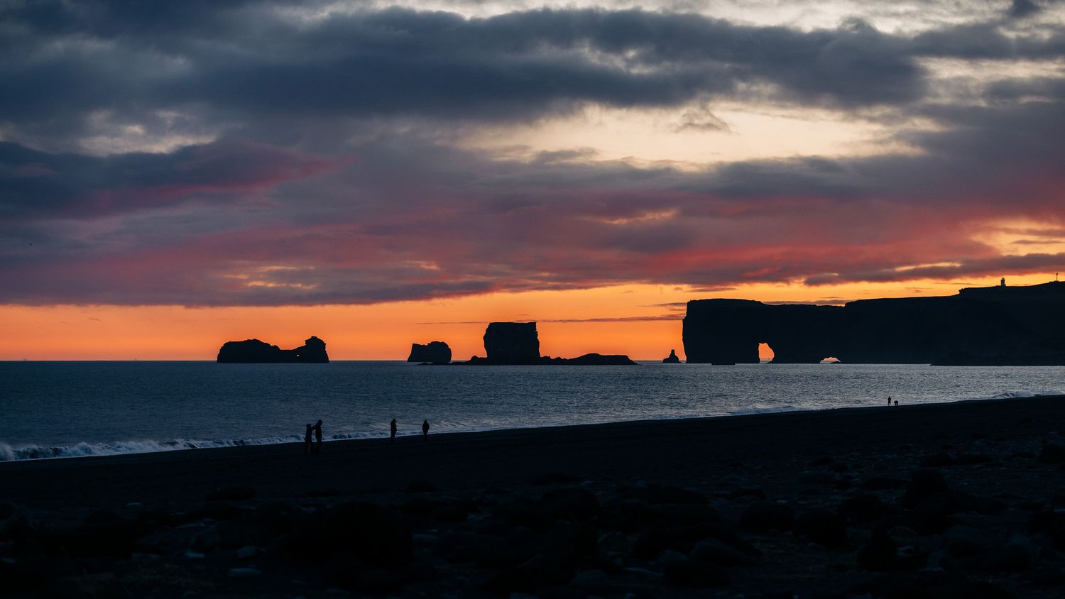Dramatic sunset silhouettes coastal rock formations against vibrant orange and purple sky over ocean horizon.