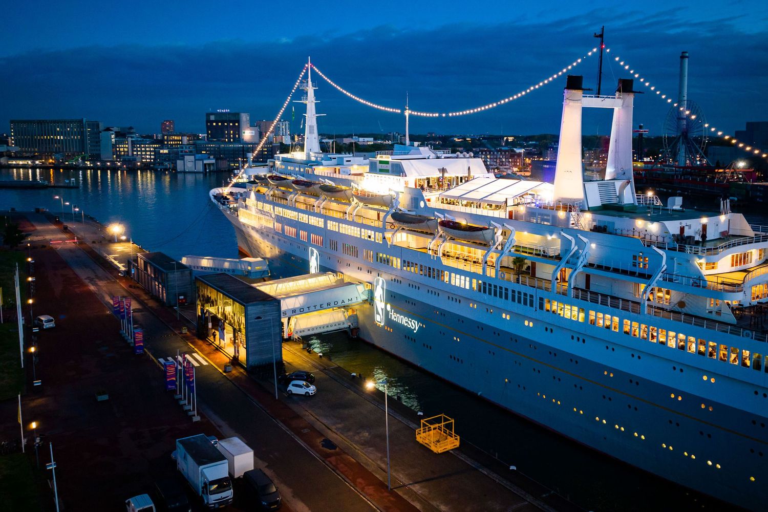Cruise ship docked at illuminated harbor at night with bridge lights reflecting on water.