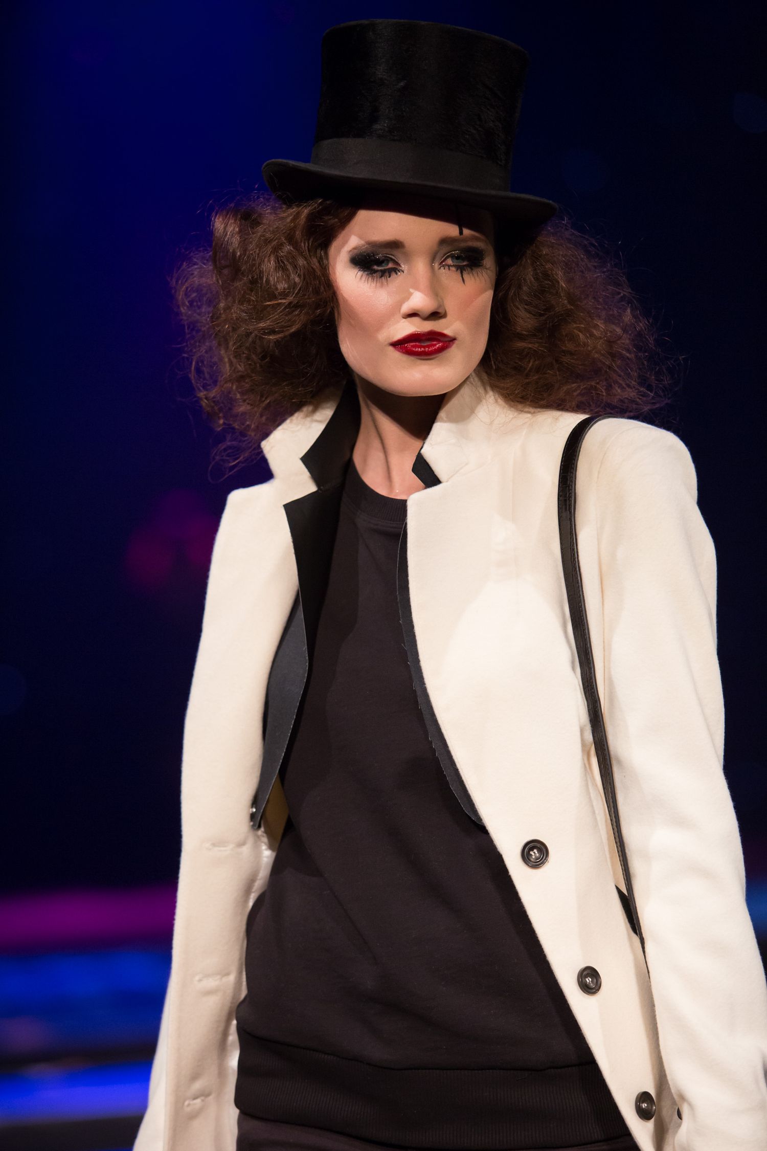Female model on runway wearing white blazer, black outfit, and top hat with dramatic makeup.