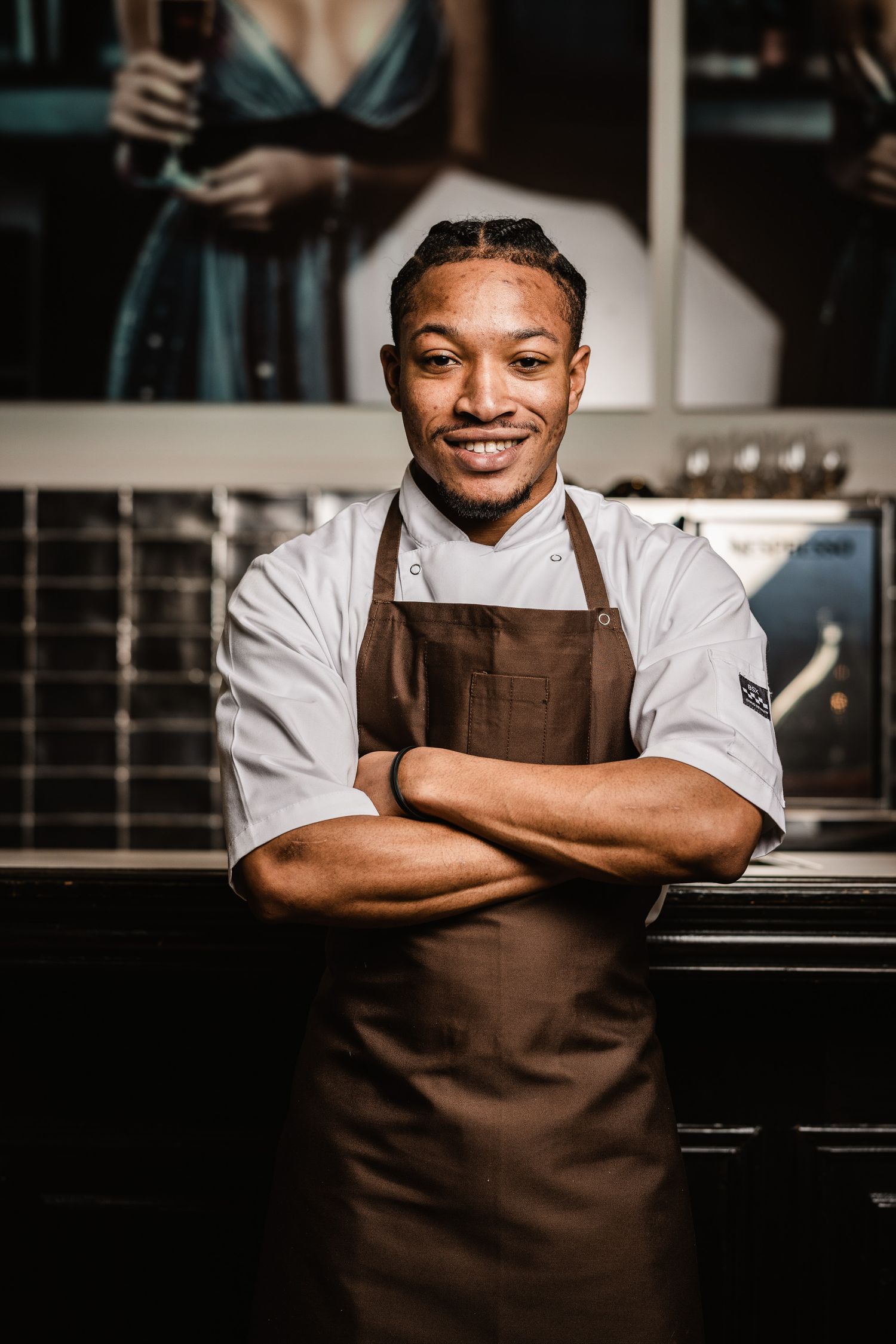 Confident chef in white coat and brown apron stands with crossed arms in professional kitchen.