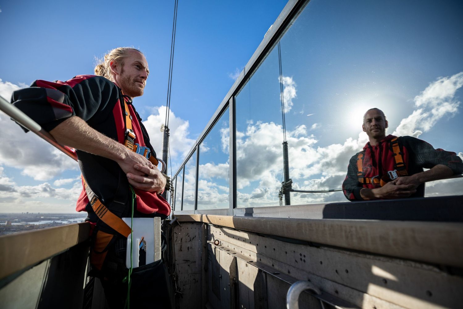 Two men in red and black jerseys stand on a rooftop observation deck under a bright sun with cloudy skies.